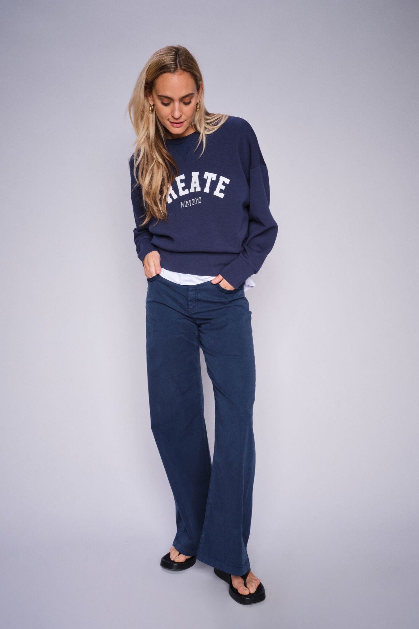 Woman wearing a navy sweatshirt with 'CREATE' text on front and navy wide leg jeans with sandals on a plain background