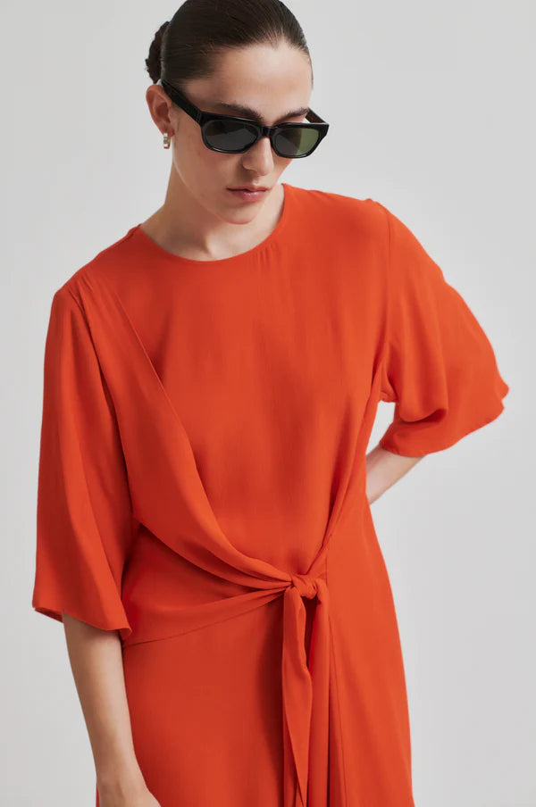 Model wearing orange short sleeve midi dress with asymmetrical hem and tie waist. Wearing black boots and sunglasses.