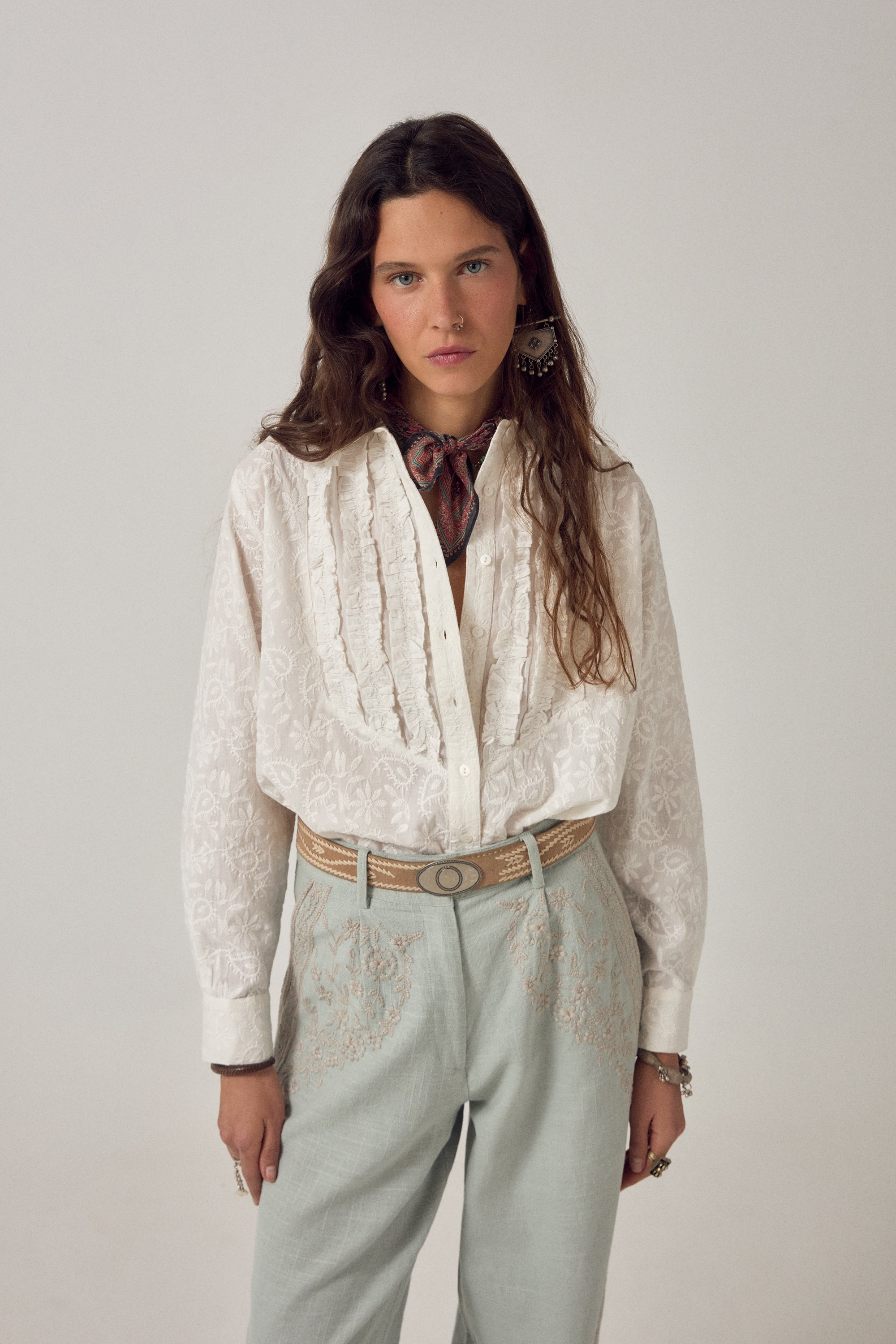 Woman wearing ecru long sleeve embroidered shirt with frill detail on a plain background. 