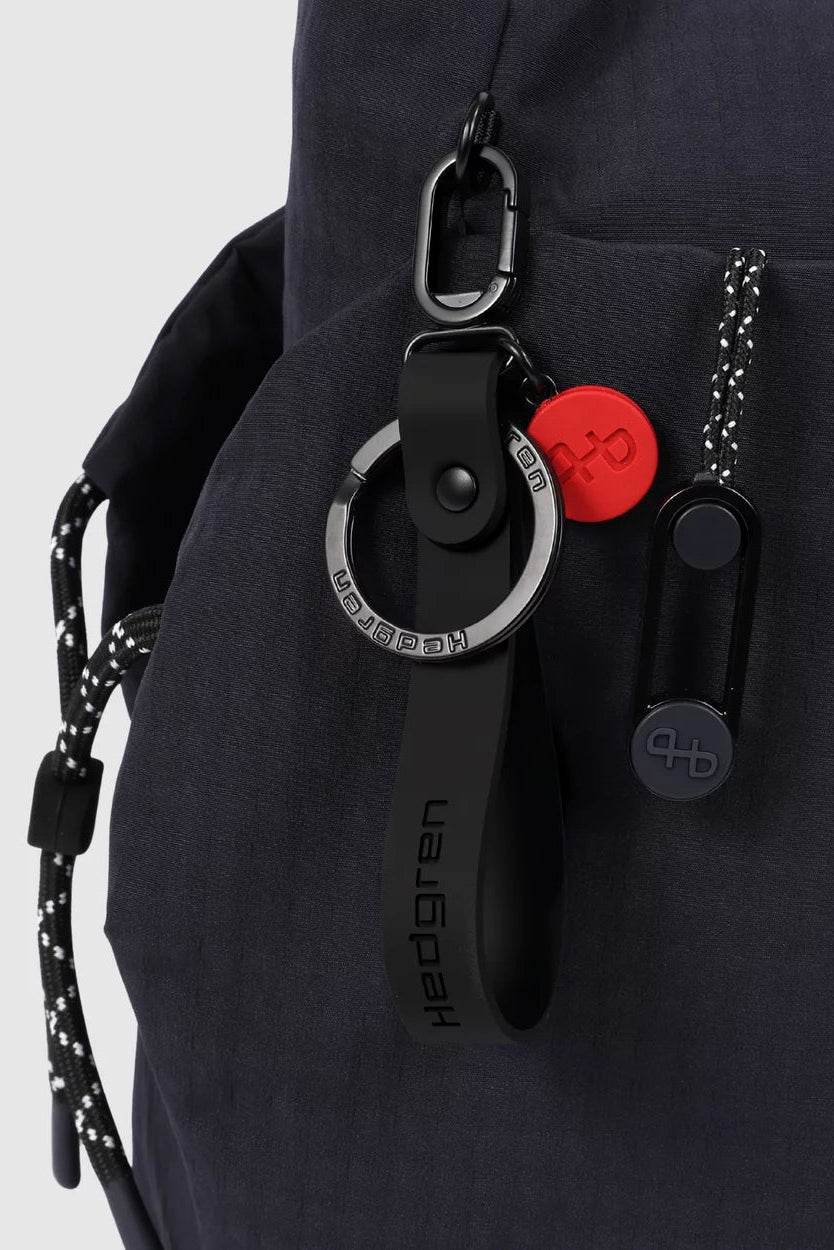 Close-up of a dark navy cross body bag with black and silver keychain-style accessories attached, including a red circular tag and a black strap with the text 'hedgren'.