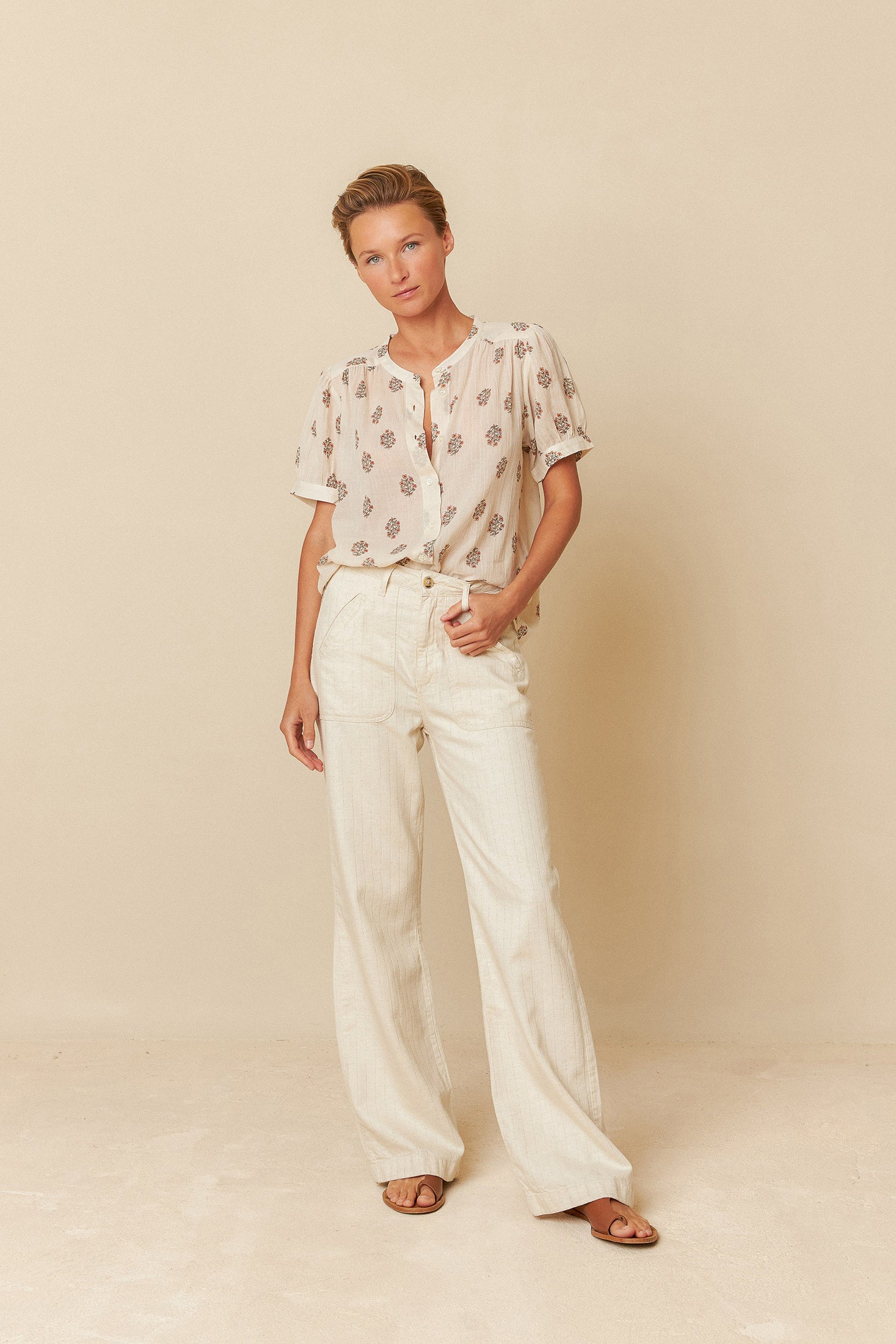 Woman wearing a white blouse with floral patterns and white pants on a beige background