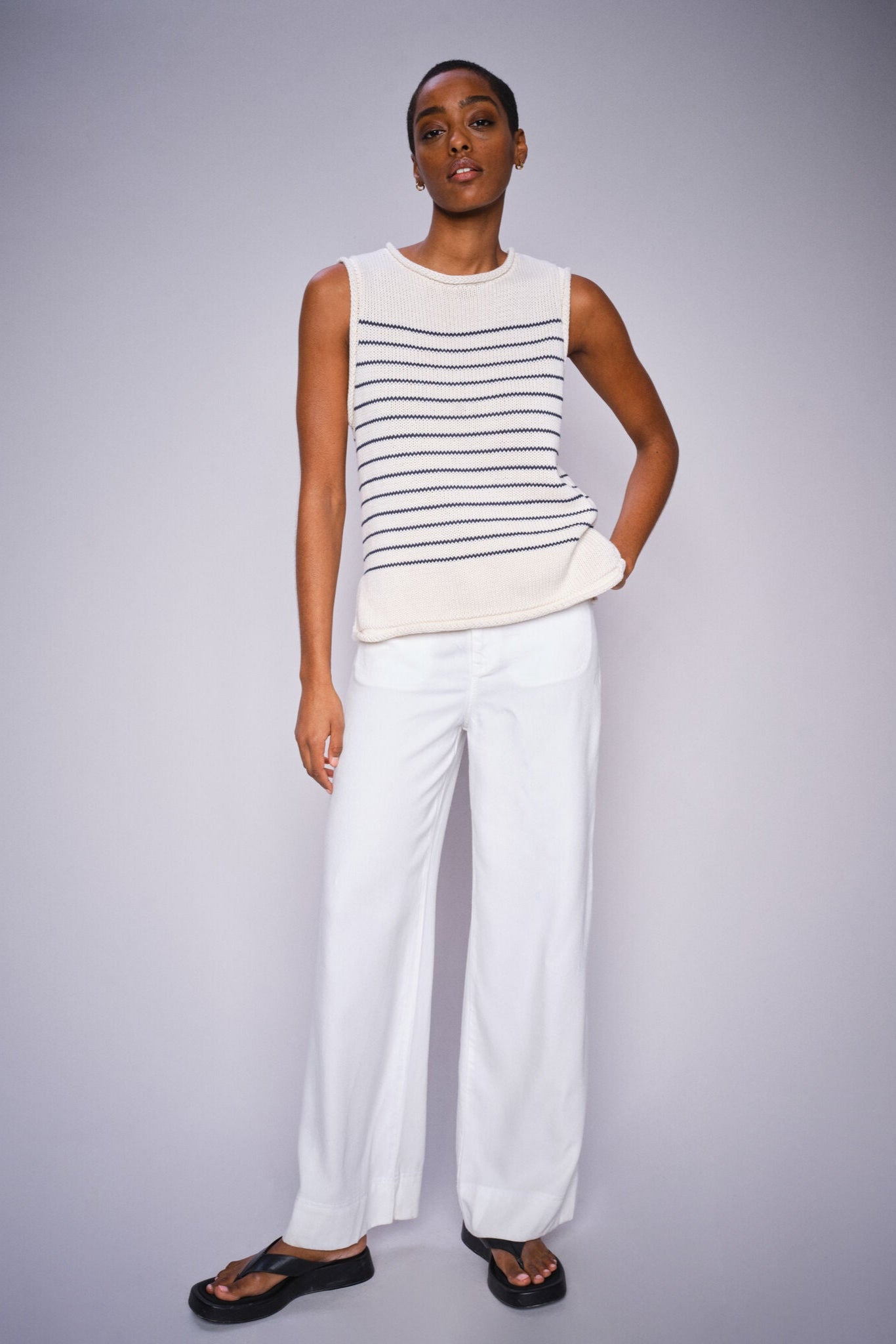 Woman wearing a striped sleeveless top and white pants on a gray background