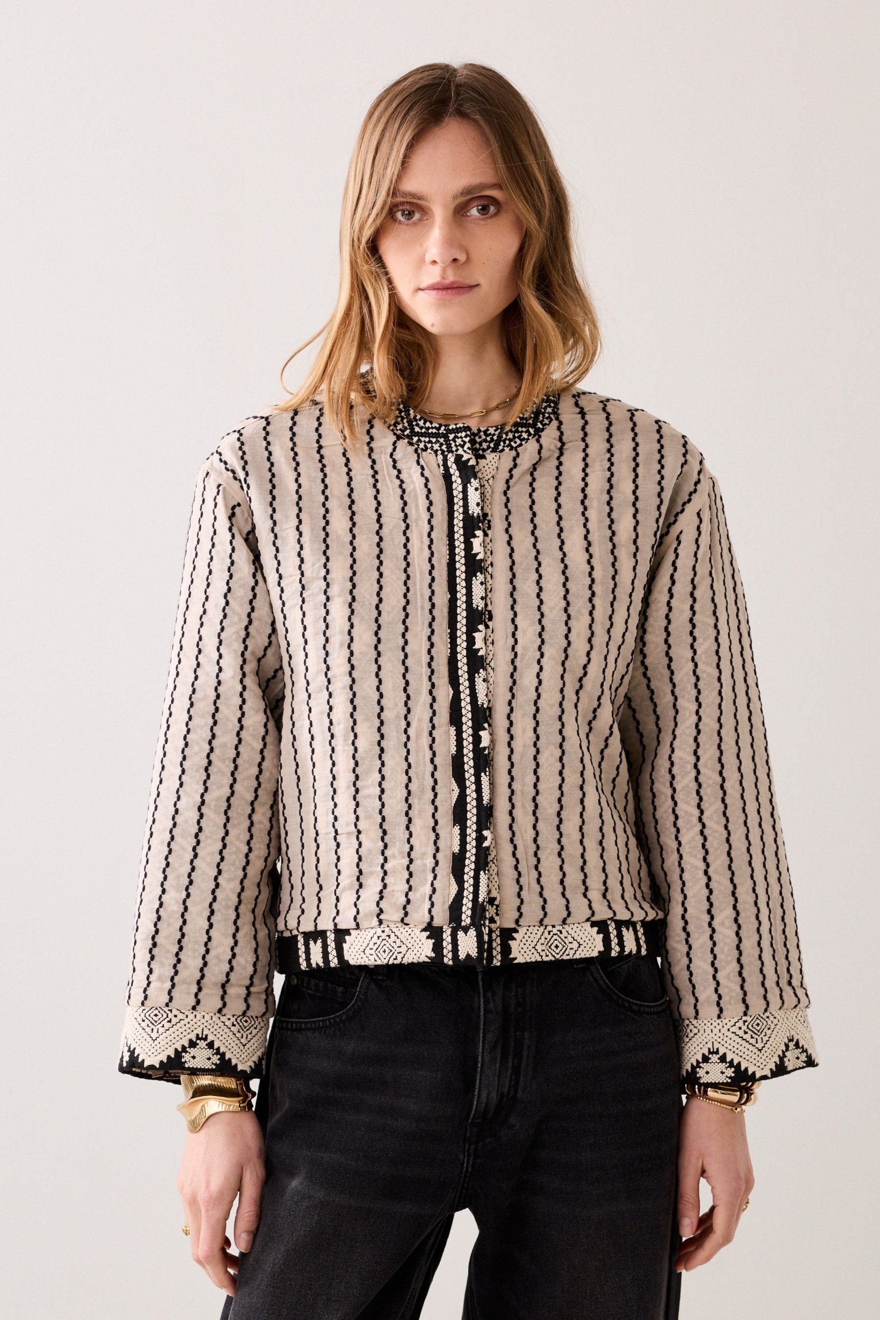 Woman wearing a patterned jacket in black and cream with long sleeves and grey jeans on a plain background