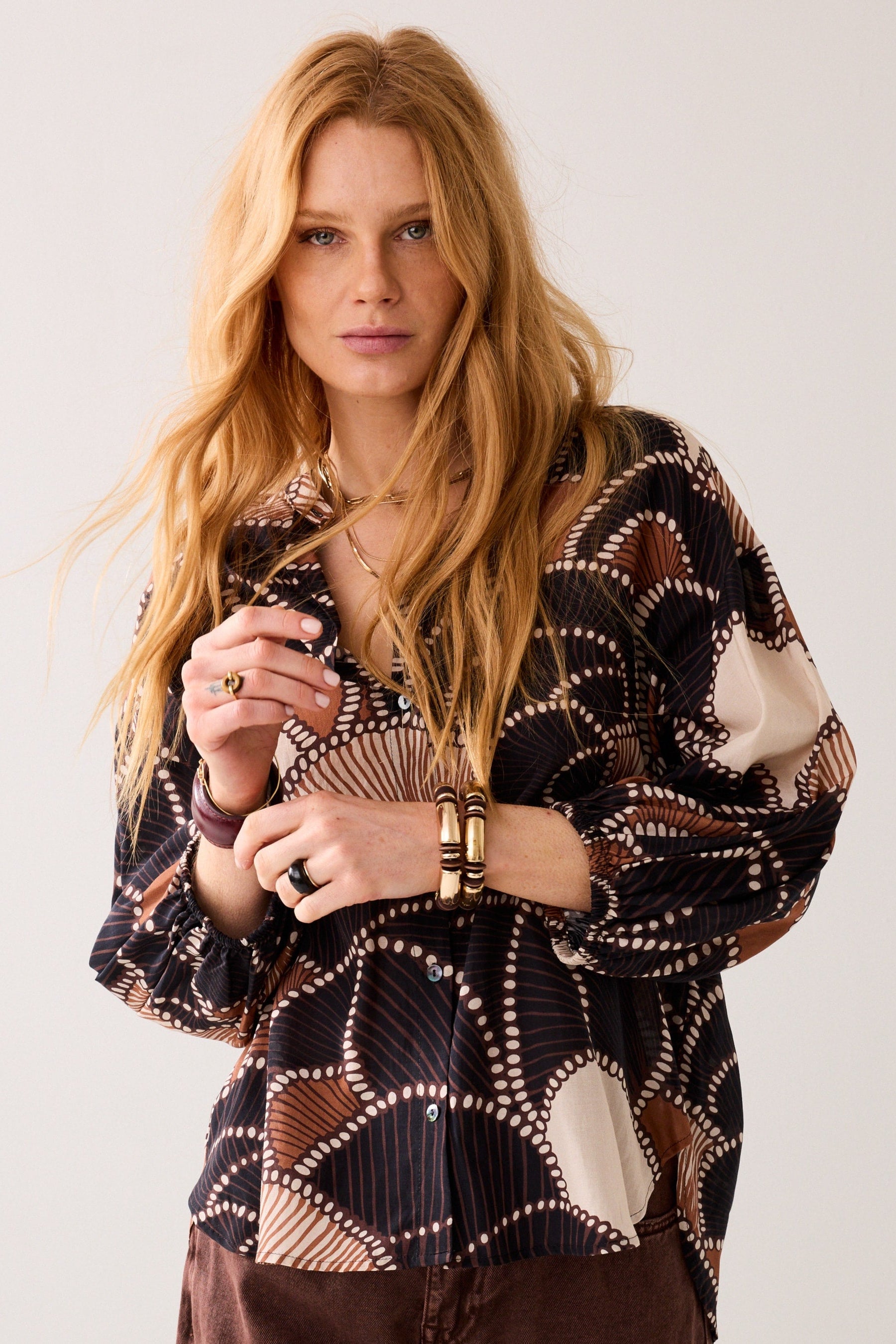 A women wearing a multi colour long-sleeve patterned shirt with brown pants on plain background