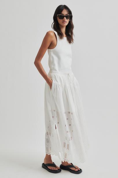 Woman wearing a white sleeveless top and white skirt with cut-out details on a plain background