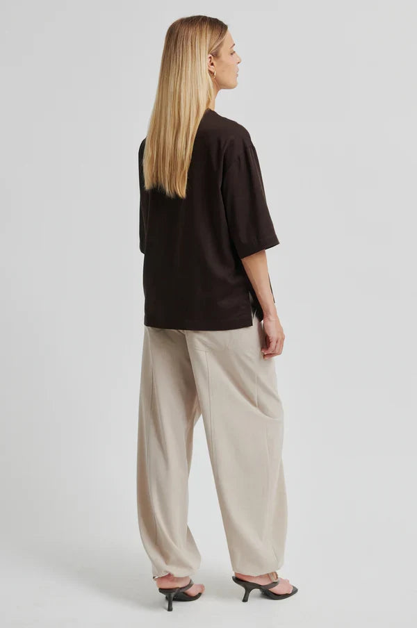 Women shown from back wearing a brown oversized shirt and beige pants on a light background