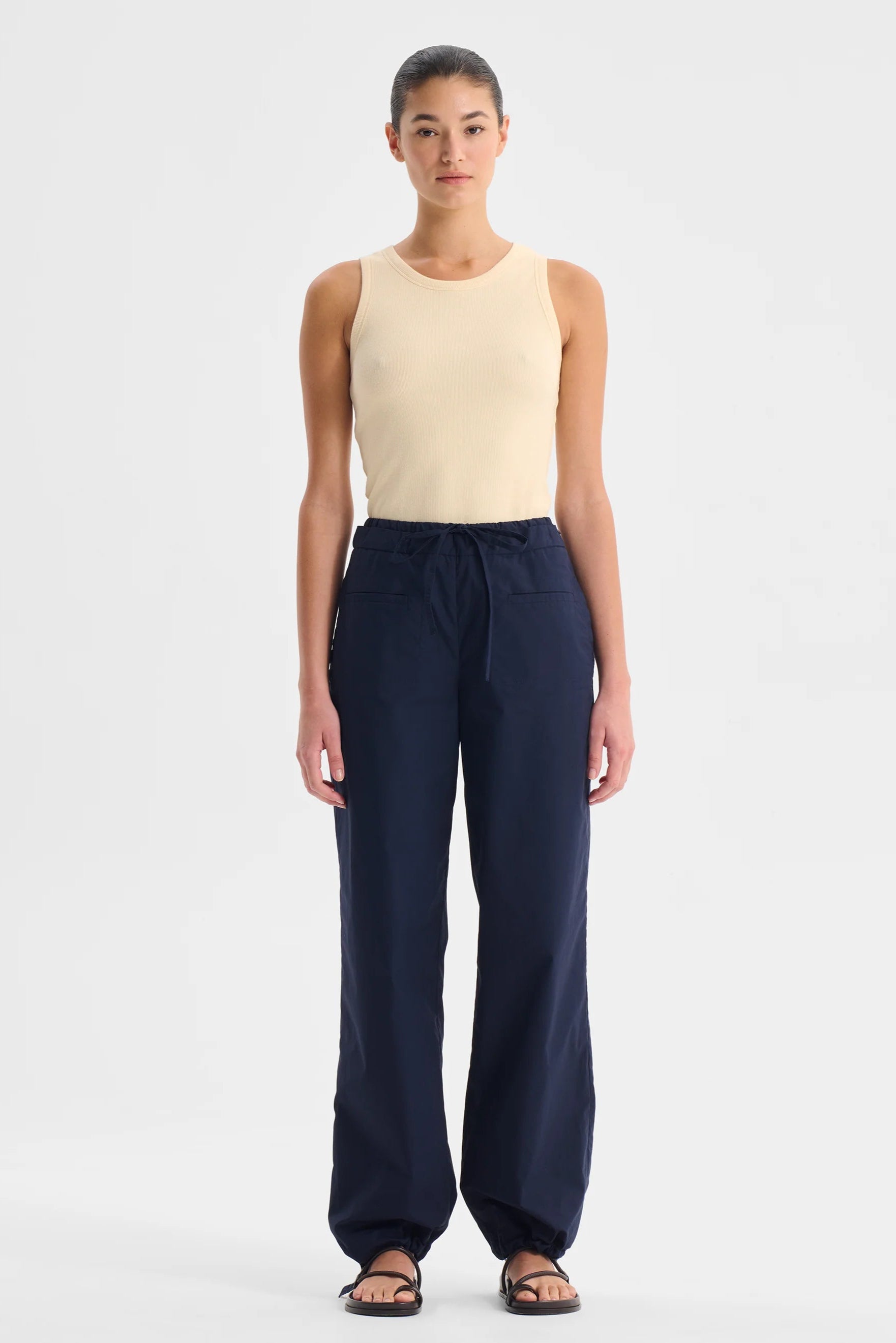 Woman wearing a cream singlet top and navy pants on a white background