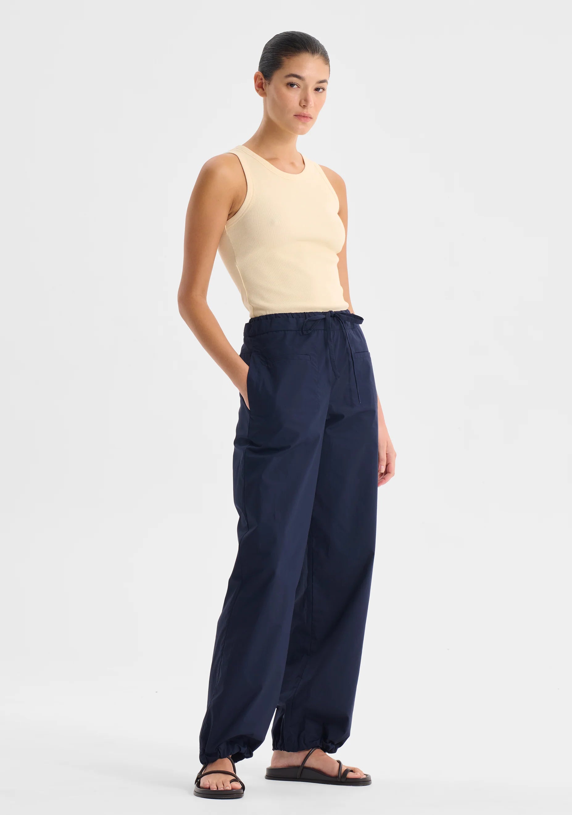 Woman wearing a cream singlet top and navy pants on a white background