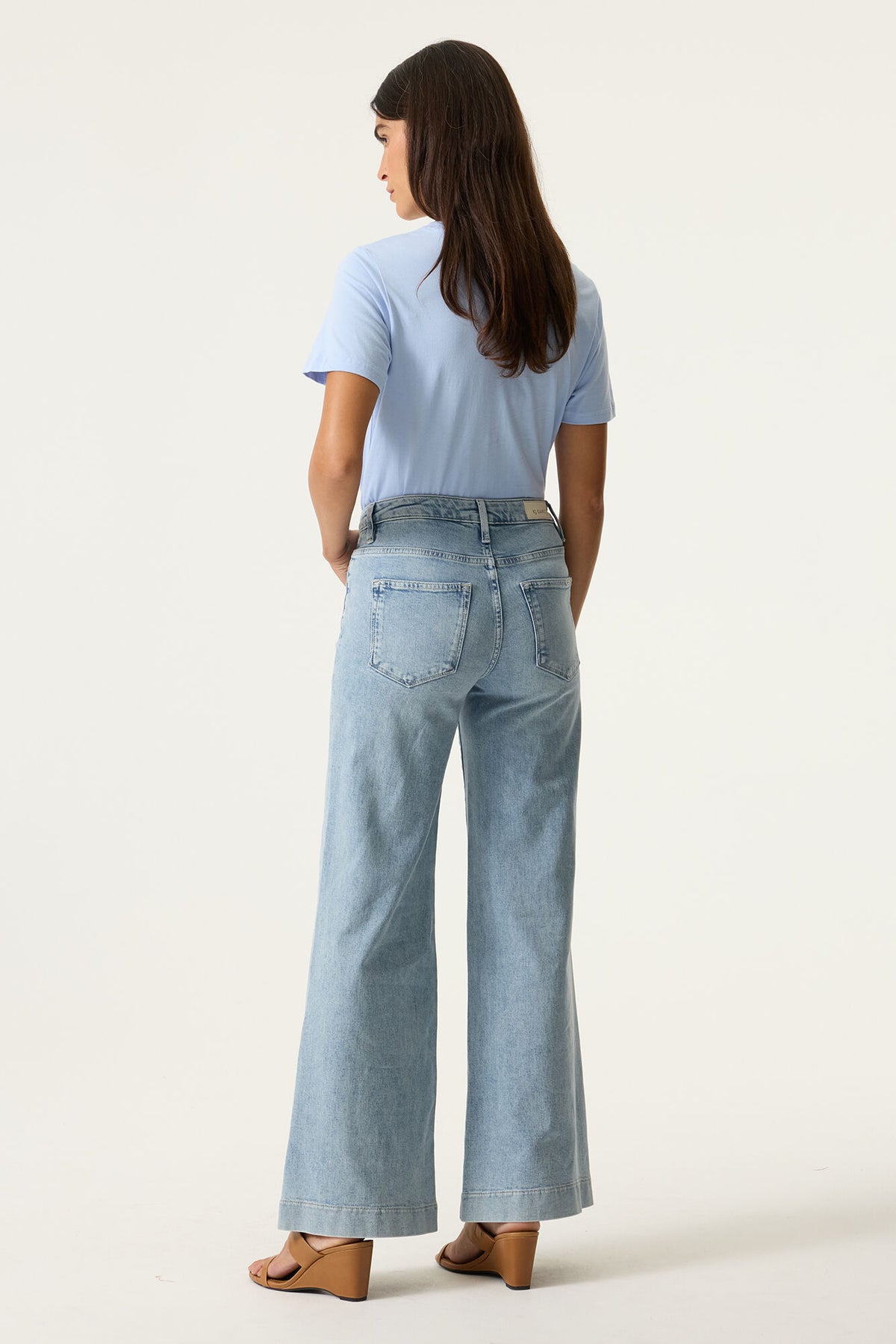 Woman shown from the back wearing light blue jeans and a matching t shirt wit brown wedge sandals on a white background