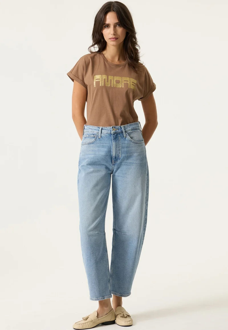 A woman modeling a brown short-sleeve graphic t-shirt with 'AMORE' printed in gold across the chest, paired with light-wash blue cropped jeans and beige loafers, against a plain white studio background.