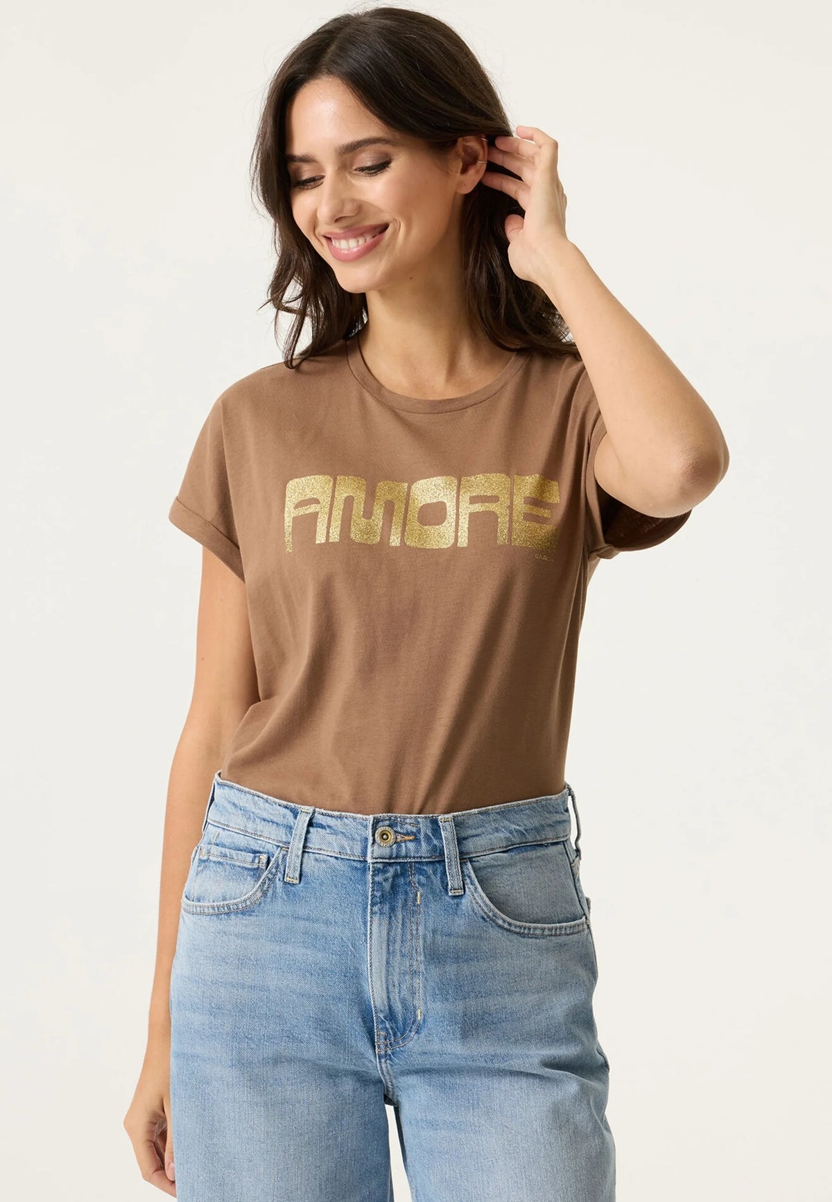 A woman wearing a brown short-sleeve t-shirt with 'AMORE' printed in gold across the chest, paired with light blue jeans, against a plain light background.