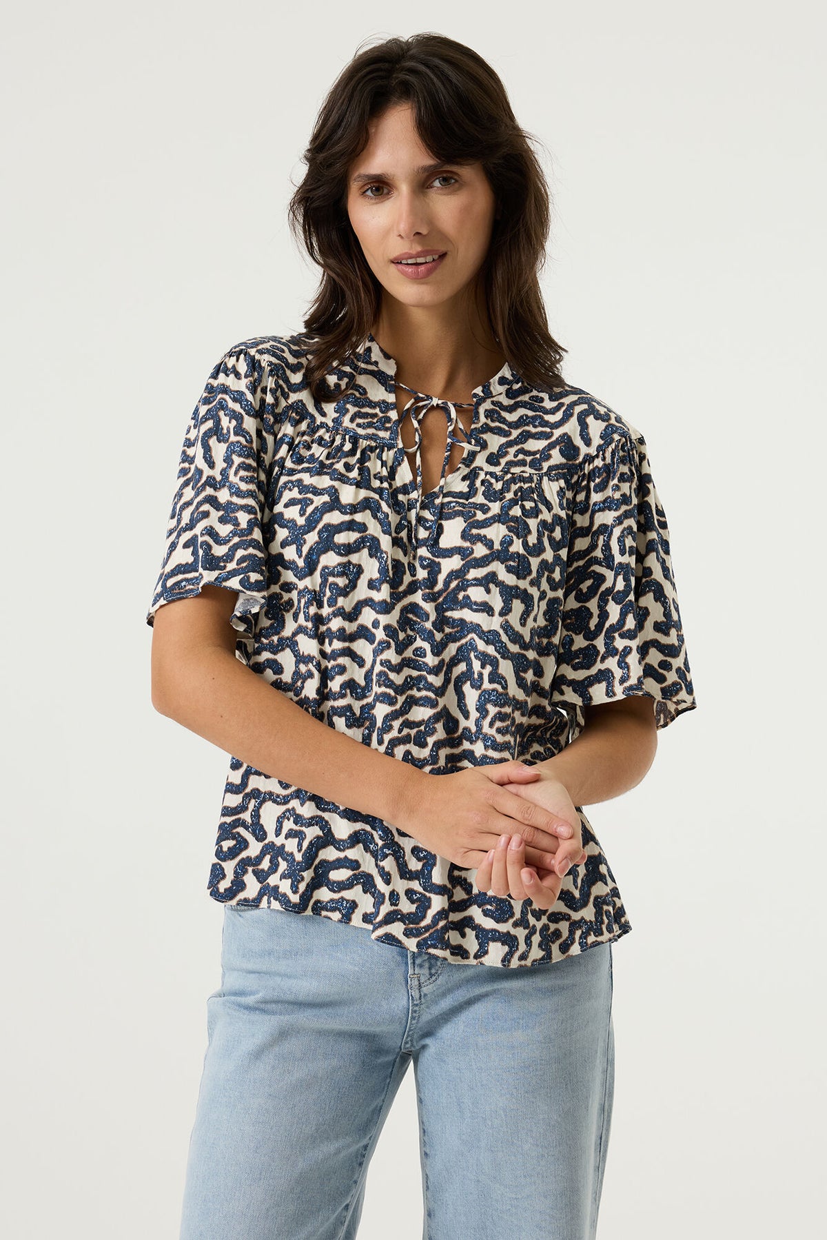 Woman wearing a patterned blouse and light blue jeans on a plain background