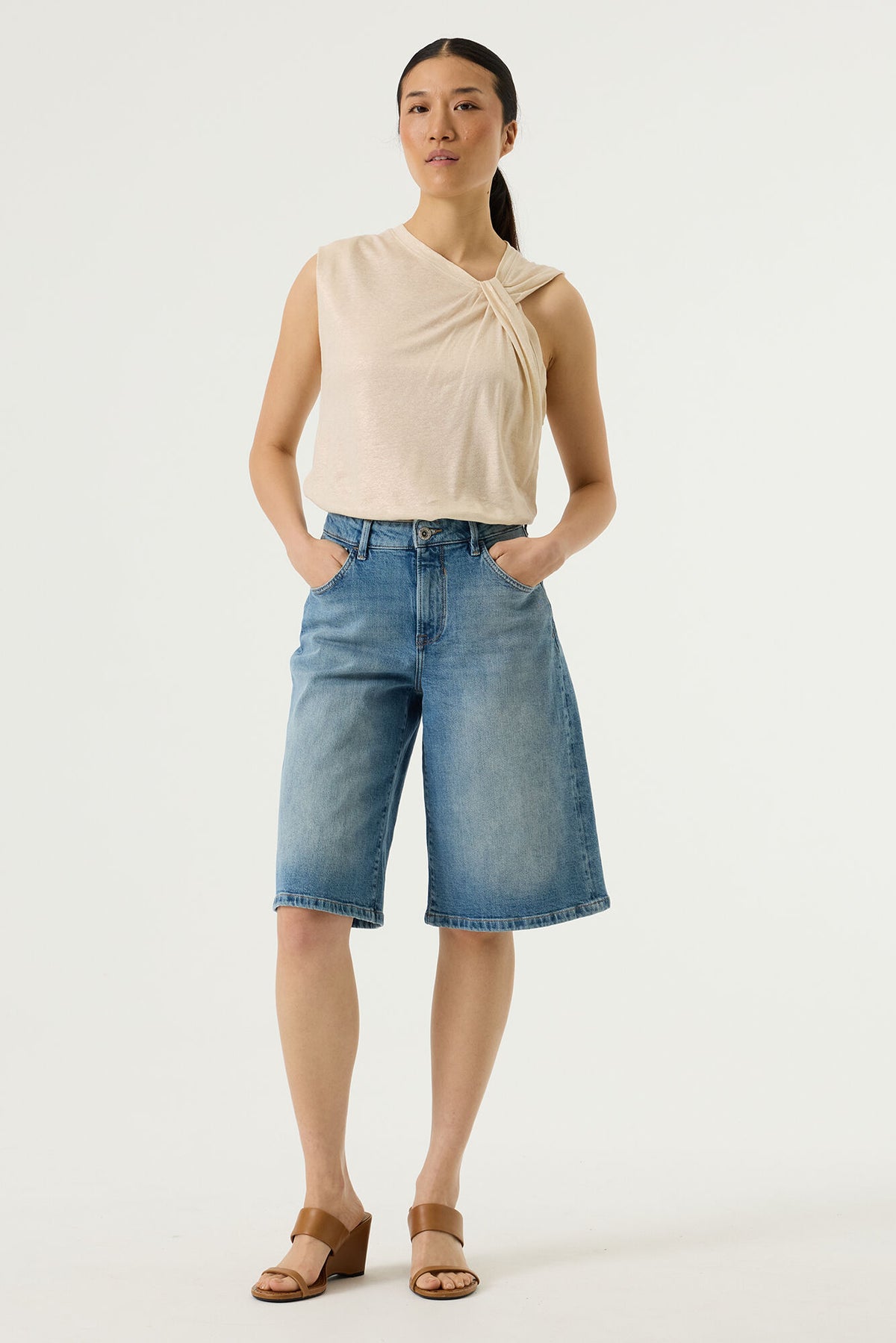 Woman wearing a beige sleeveless top and blue denim bermuda shorts on a white background
