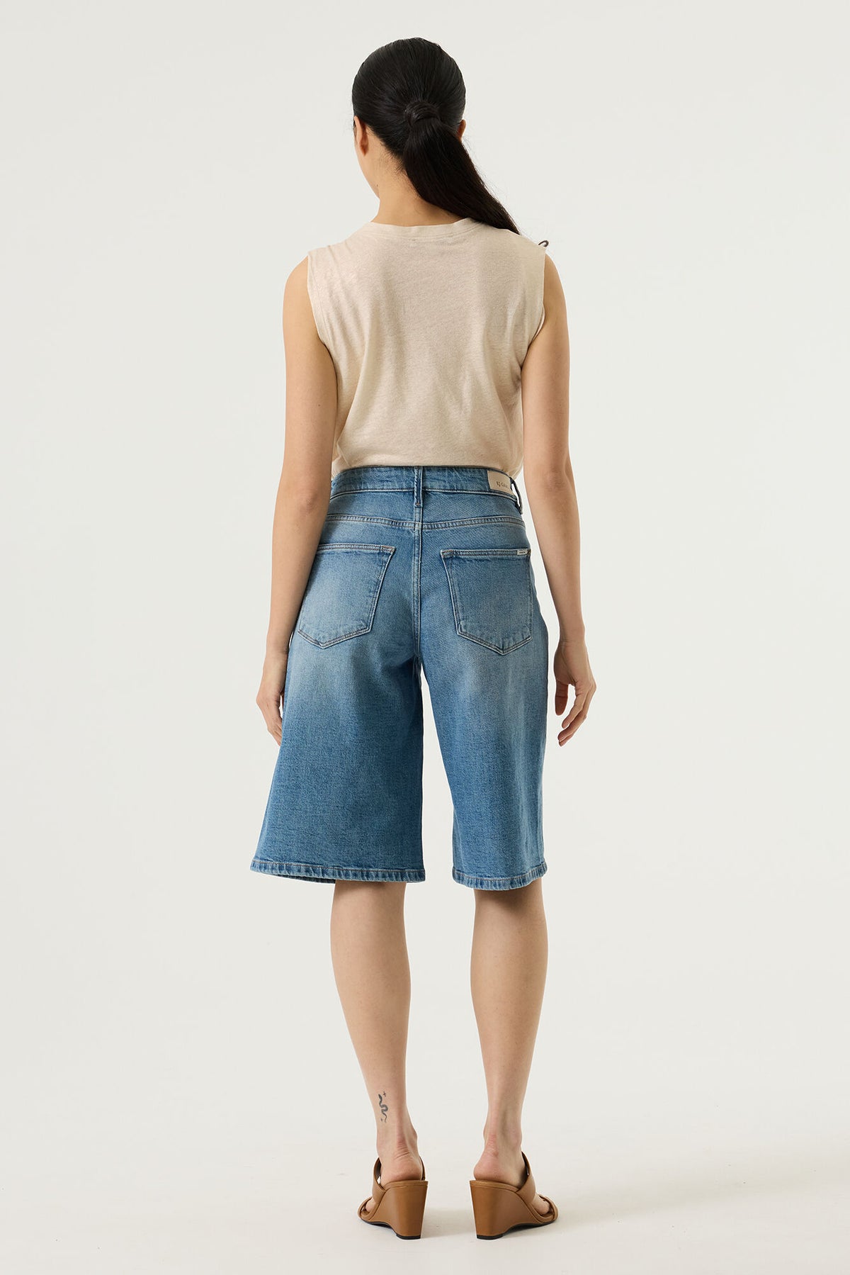 Woman shown from the back wearing a beige sleeveless top and blue denim bermuda shorts on a white background 