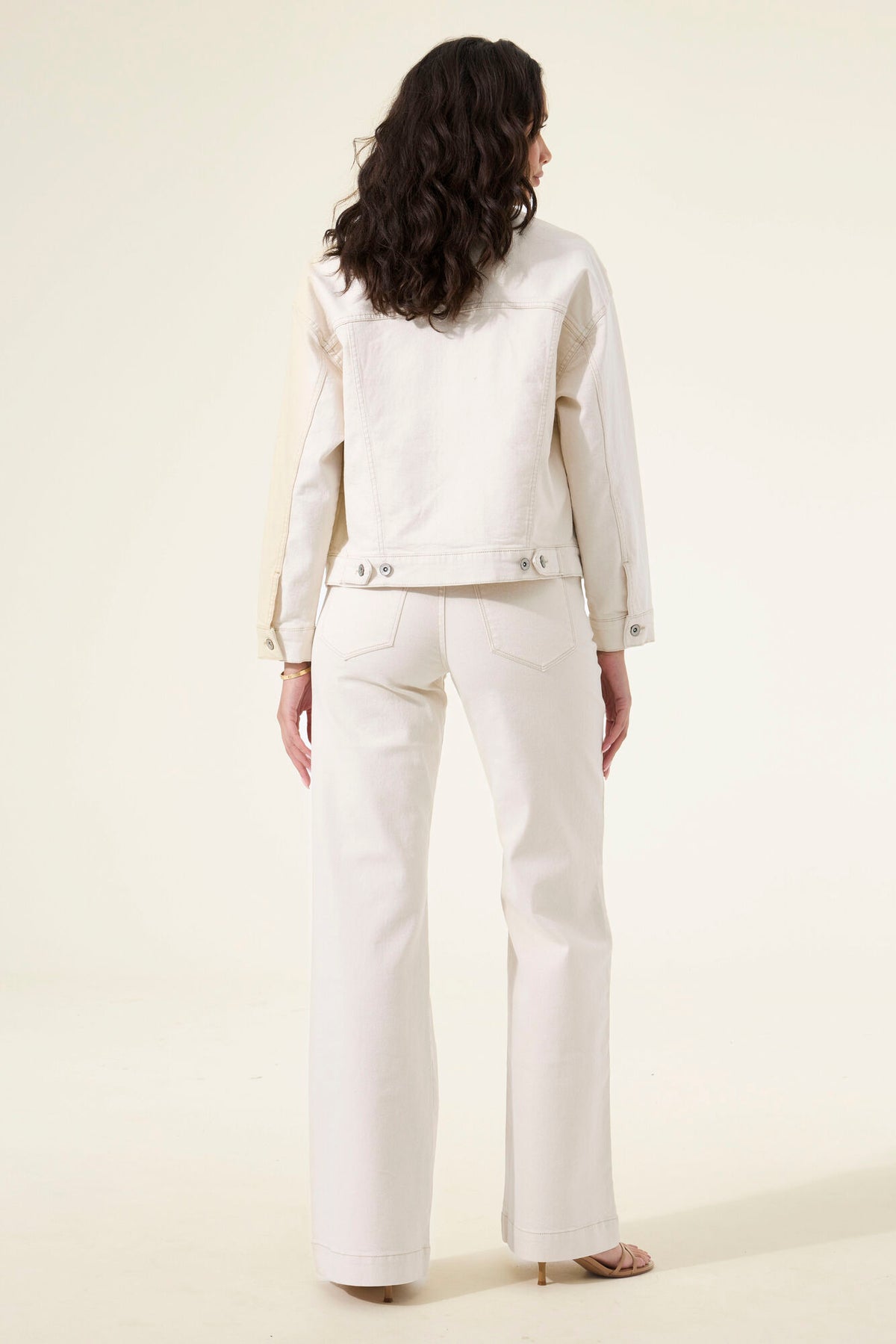 A model viewed from the back wearing a matching off-white cropped jacket and wide-leg pants set, photographed against a plain cream studio background.