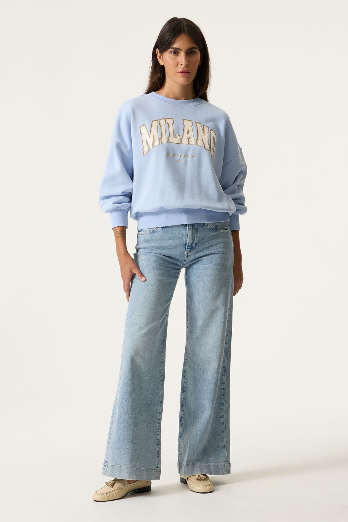 Model close up wearing a light blue sweatshirt with 'MILANO' text graphic on front and light blue jeans. 
