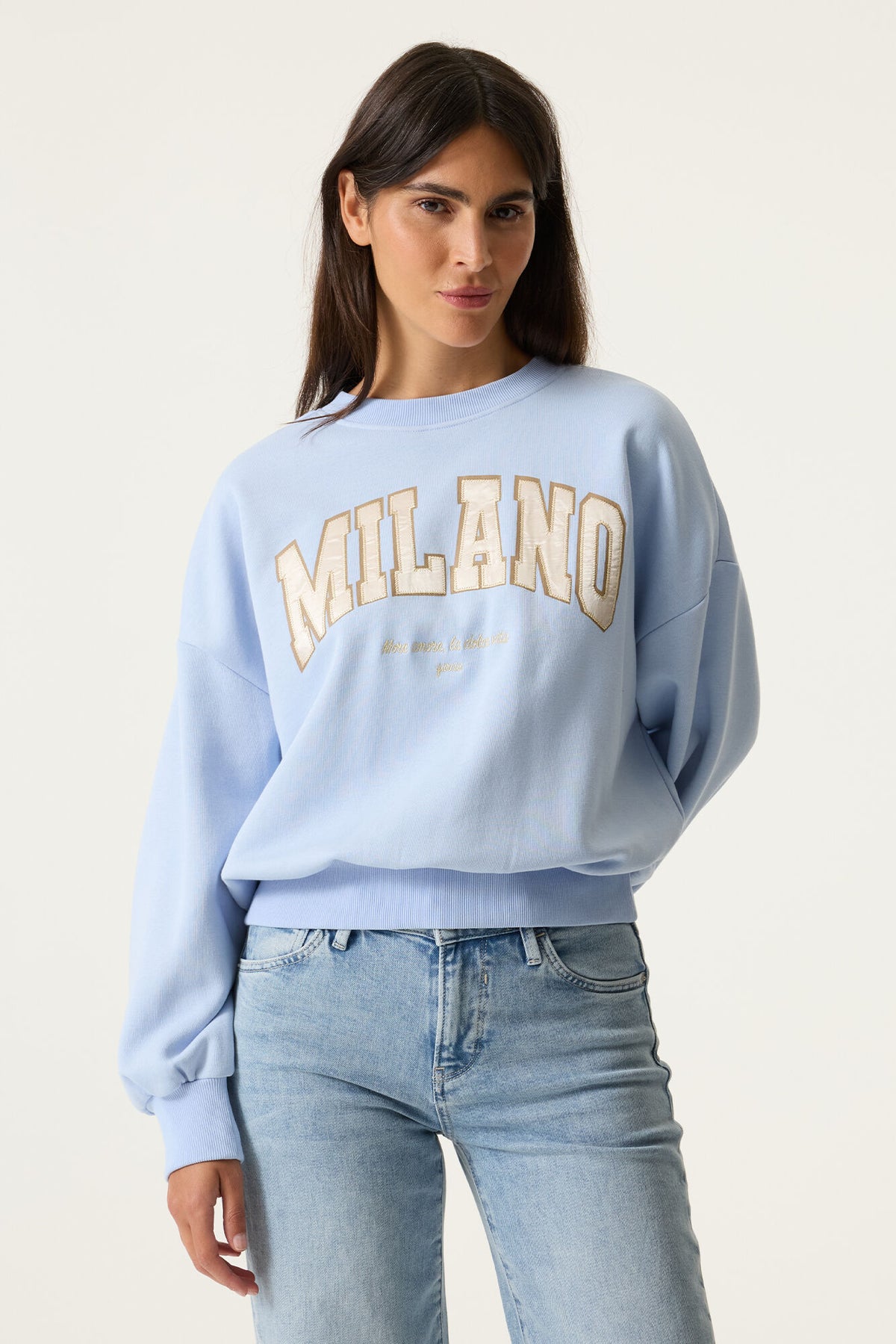 Model wearing a light blue sweatshirt with 'MILANO' text graphic on front and light blue jeans. 