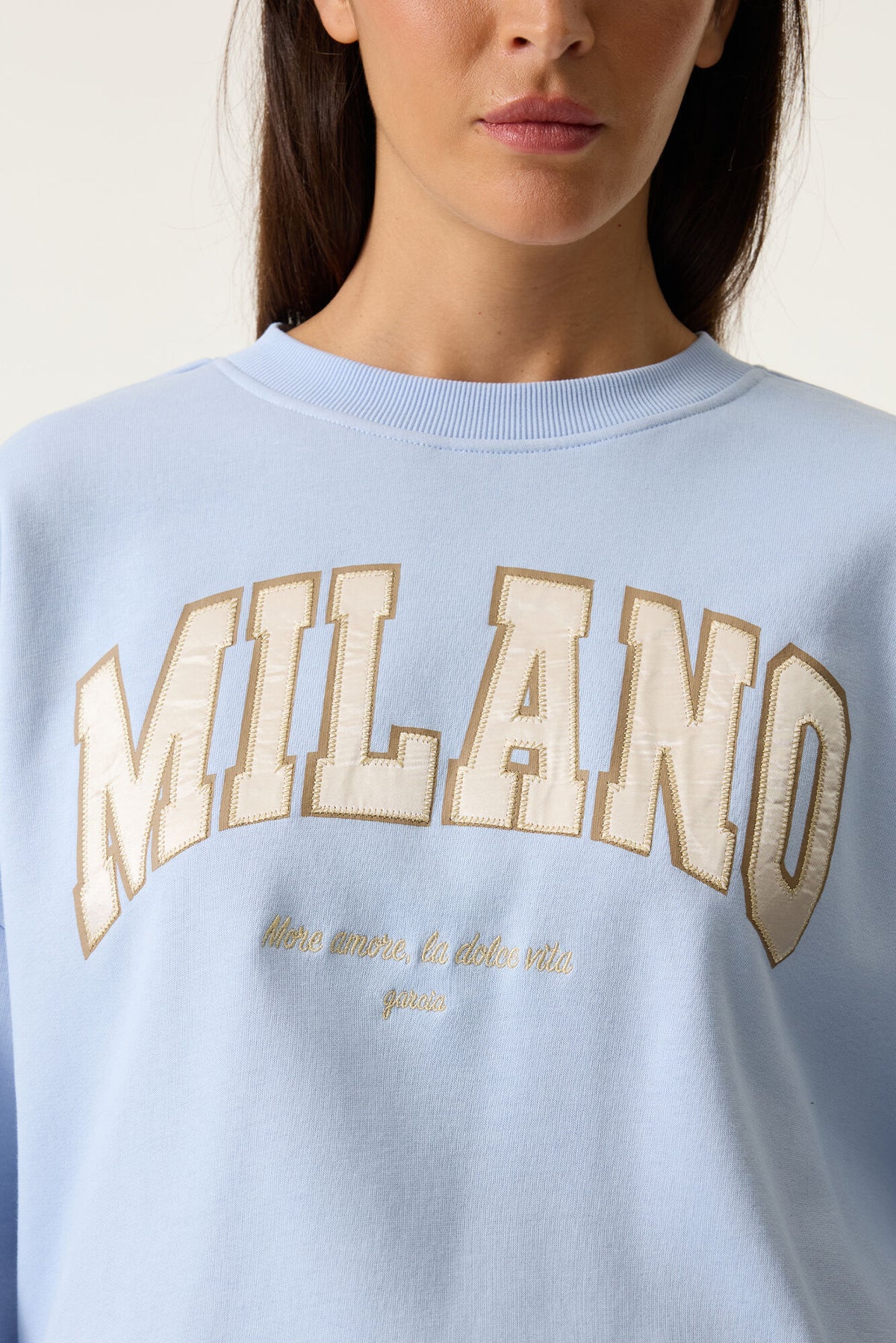 Model close up wearing a light blue sweatshirt with 'MILANO' text graphic on front 