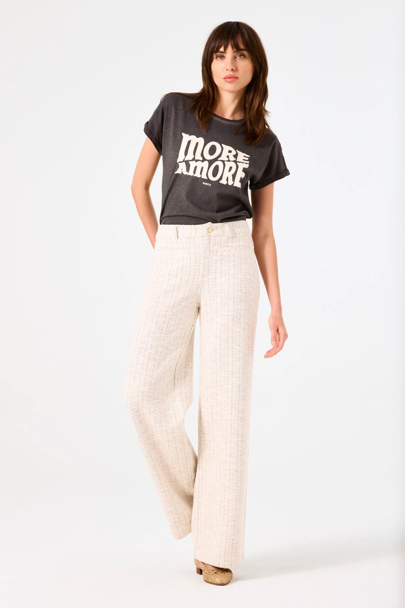 Woman wearing a dark gray t-shirt with 'MORE AMORE GARCIA' text and natural pants on a white background
