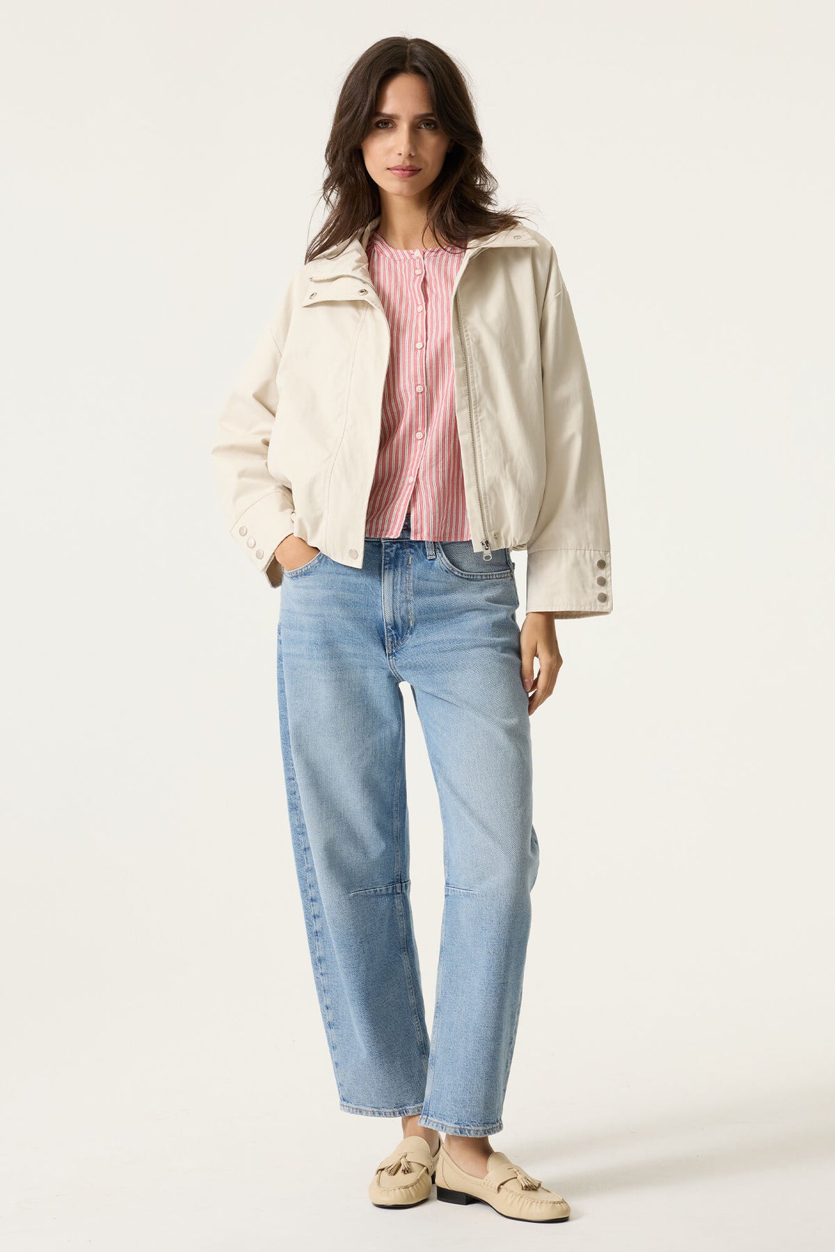 Woman wearing a cream jacket, pink striped shirt, and light blue jeans on a white background