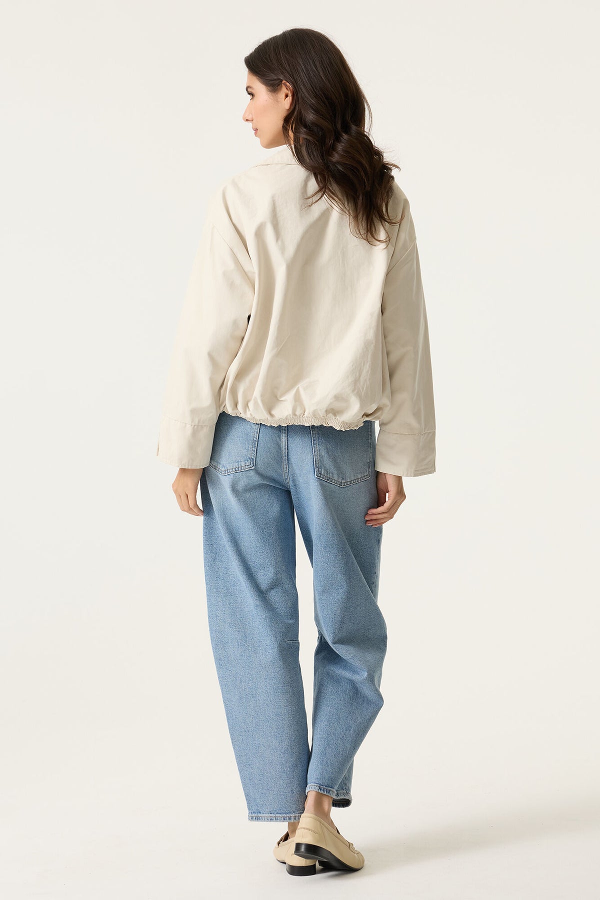 Woman wearing a cropped off white jacket and light blue denim jeans and loafers on a plain background. 