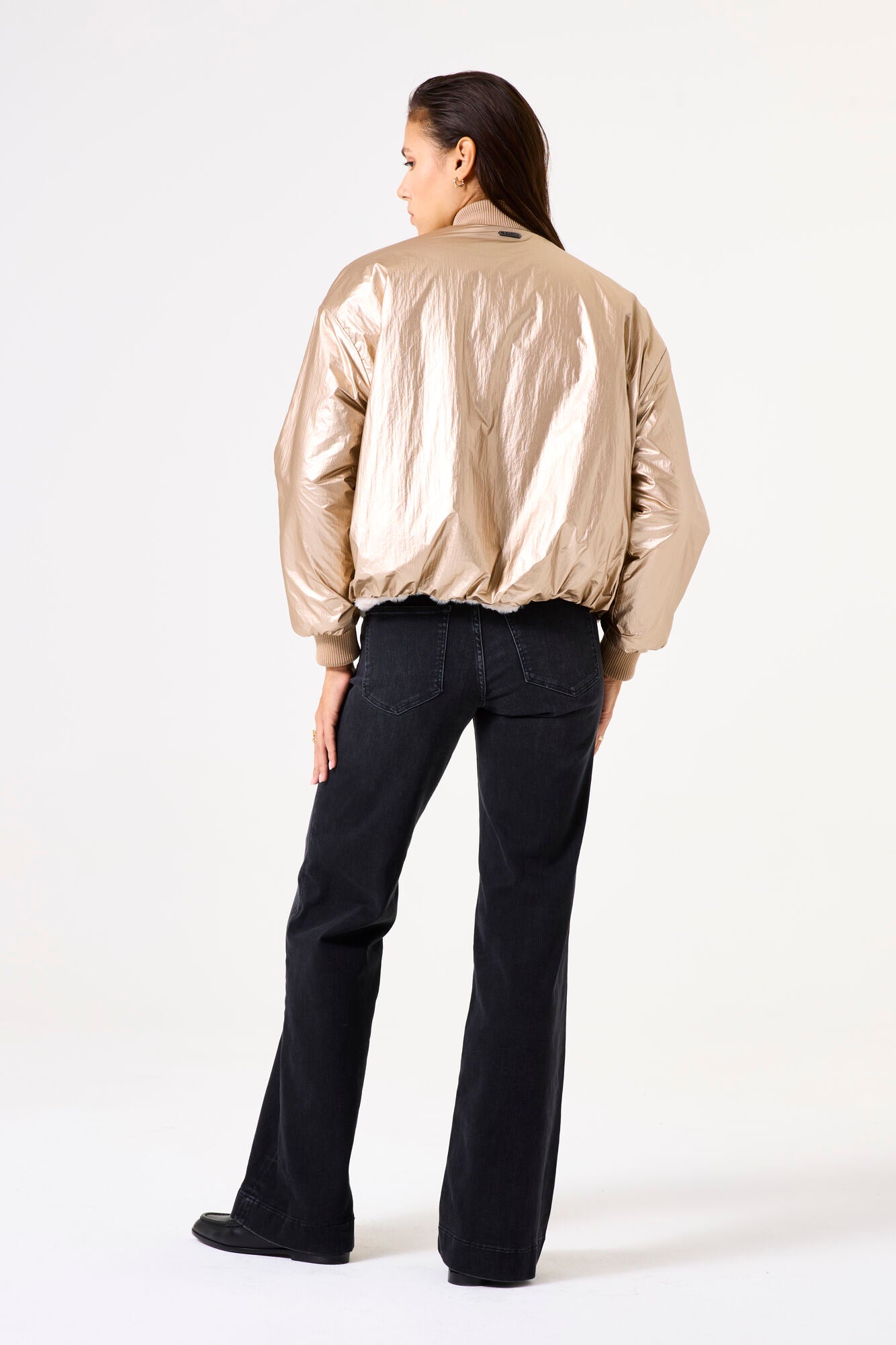 Woman wearing a gold bomber jacket and dark grey jeans s on a white background
