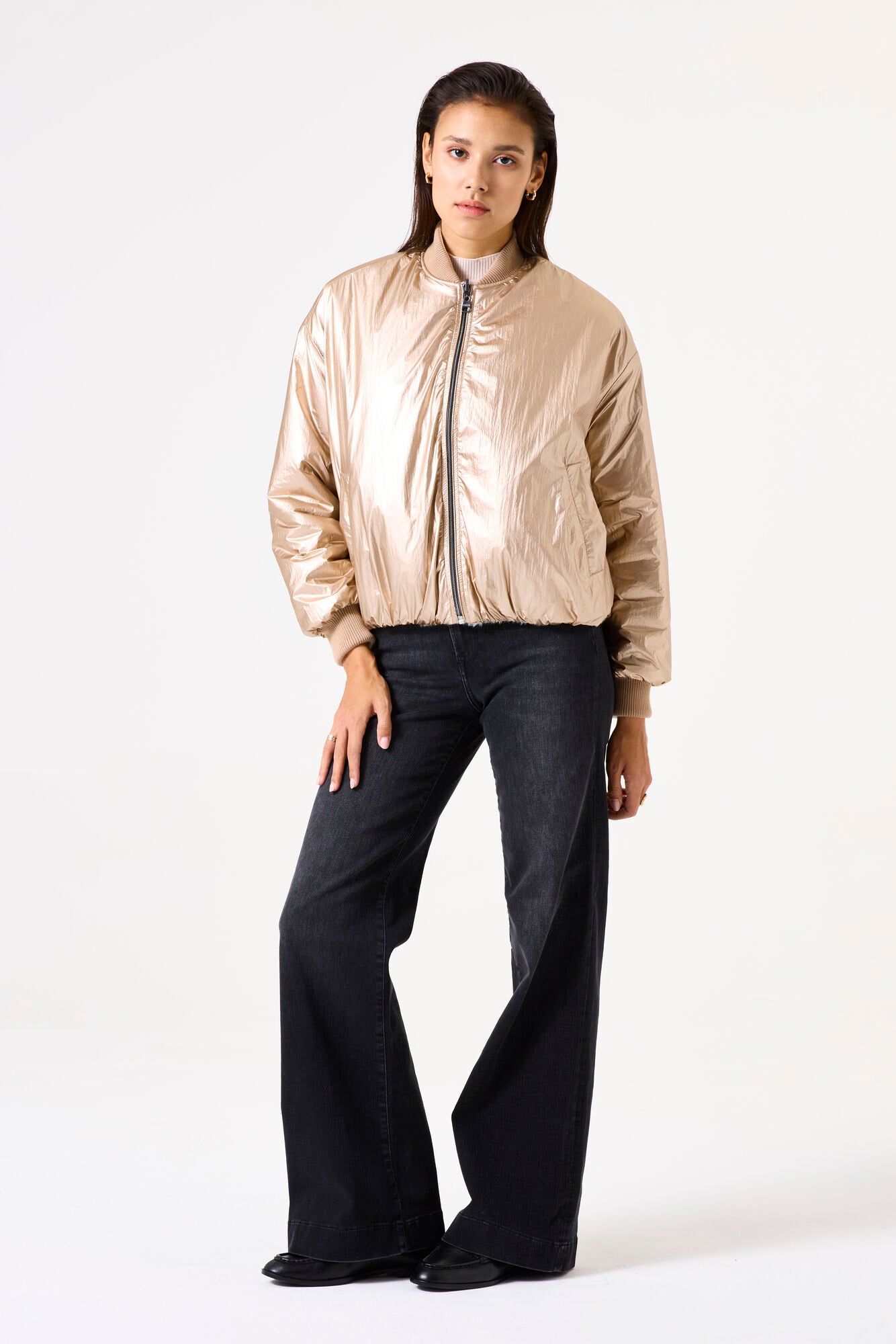 Woman wearing a gold bomber jacket and dark grey jeans s on a white background