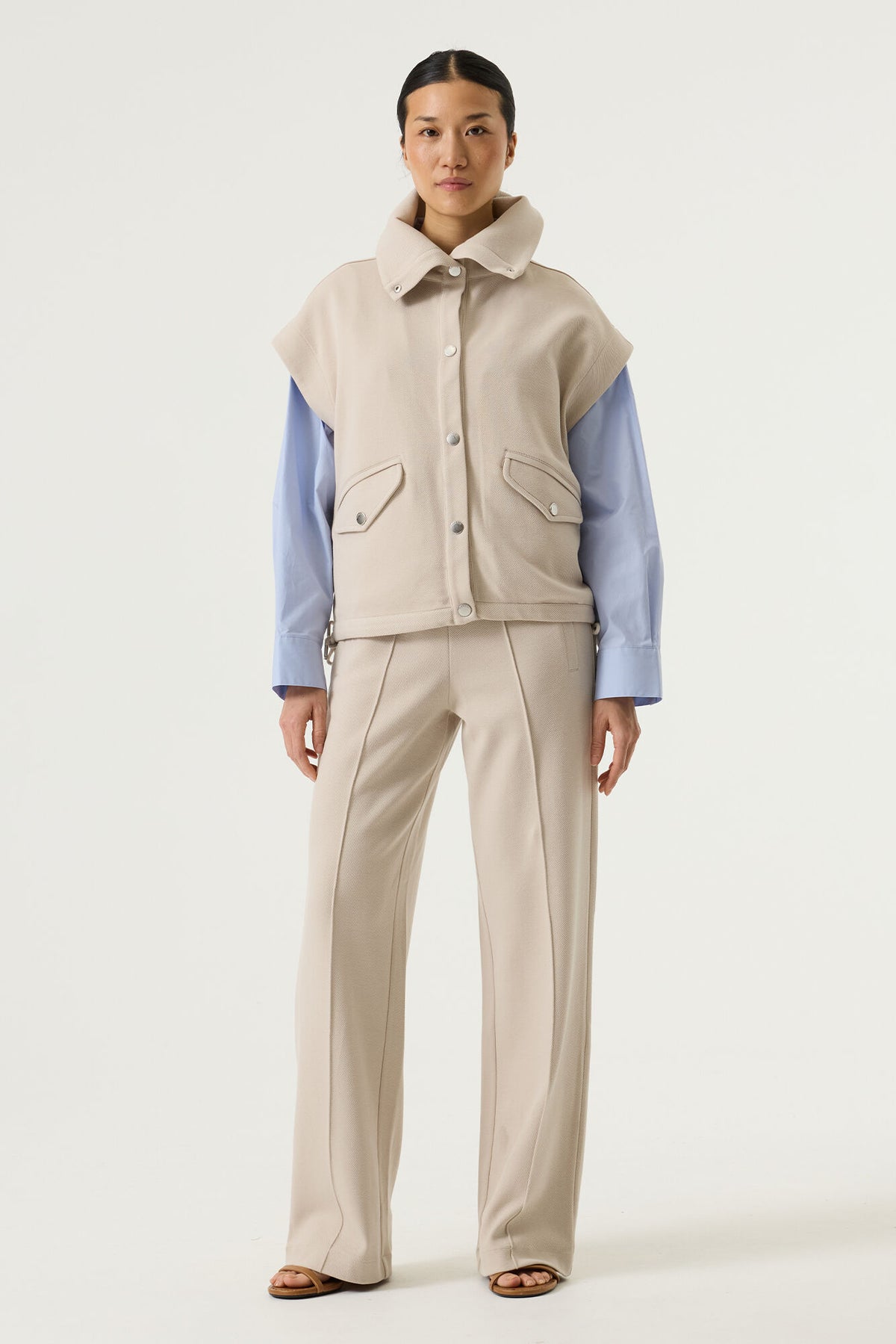 woman wearing a beige vest over a light blue shirt with beige pants on a white background
