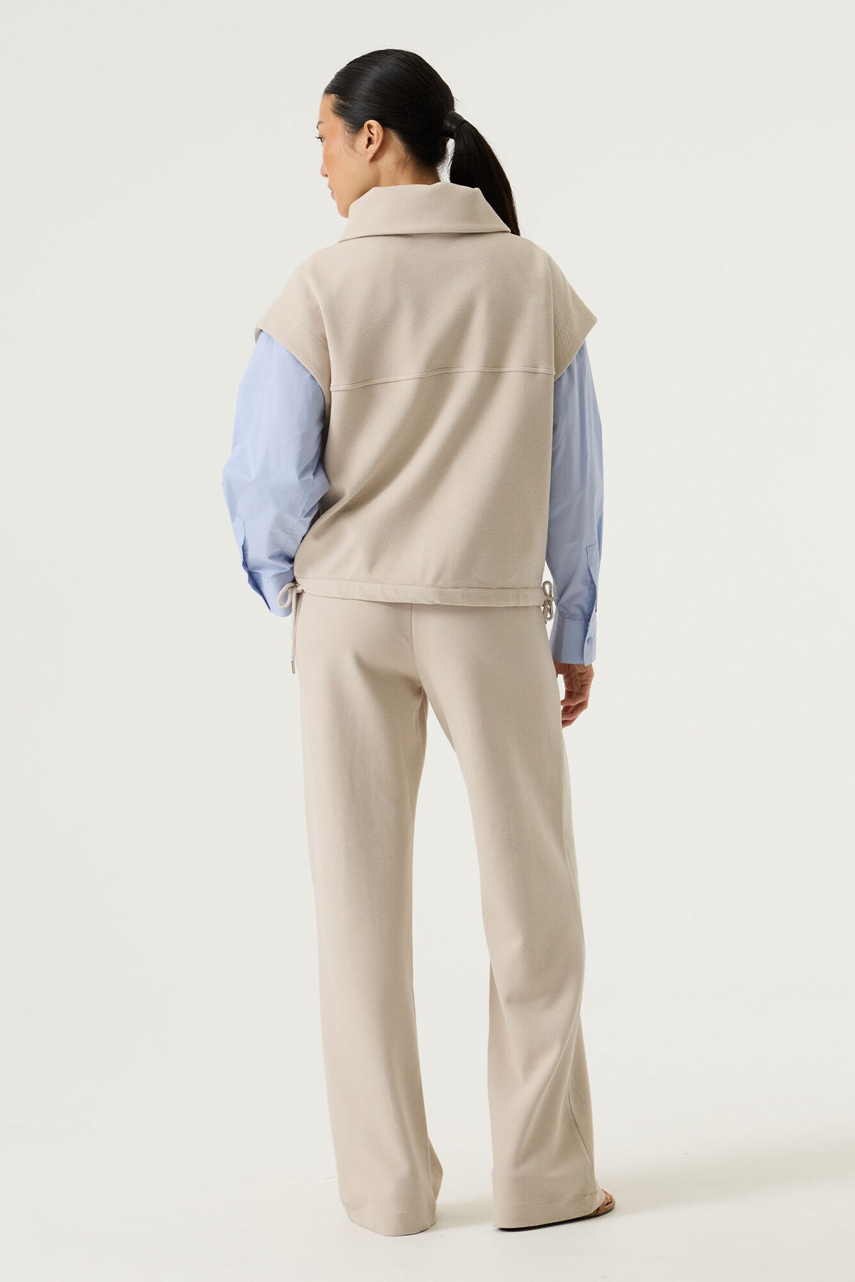 woman shown from the back wearing a beige vest over a light blue shirt with beige pants on a white background