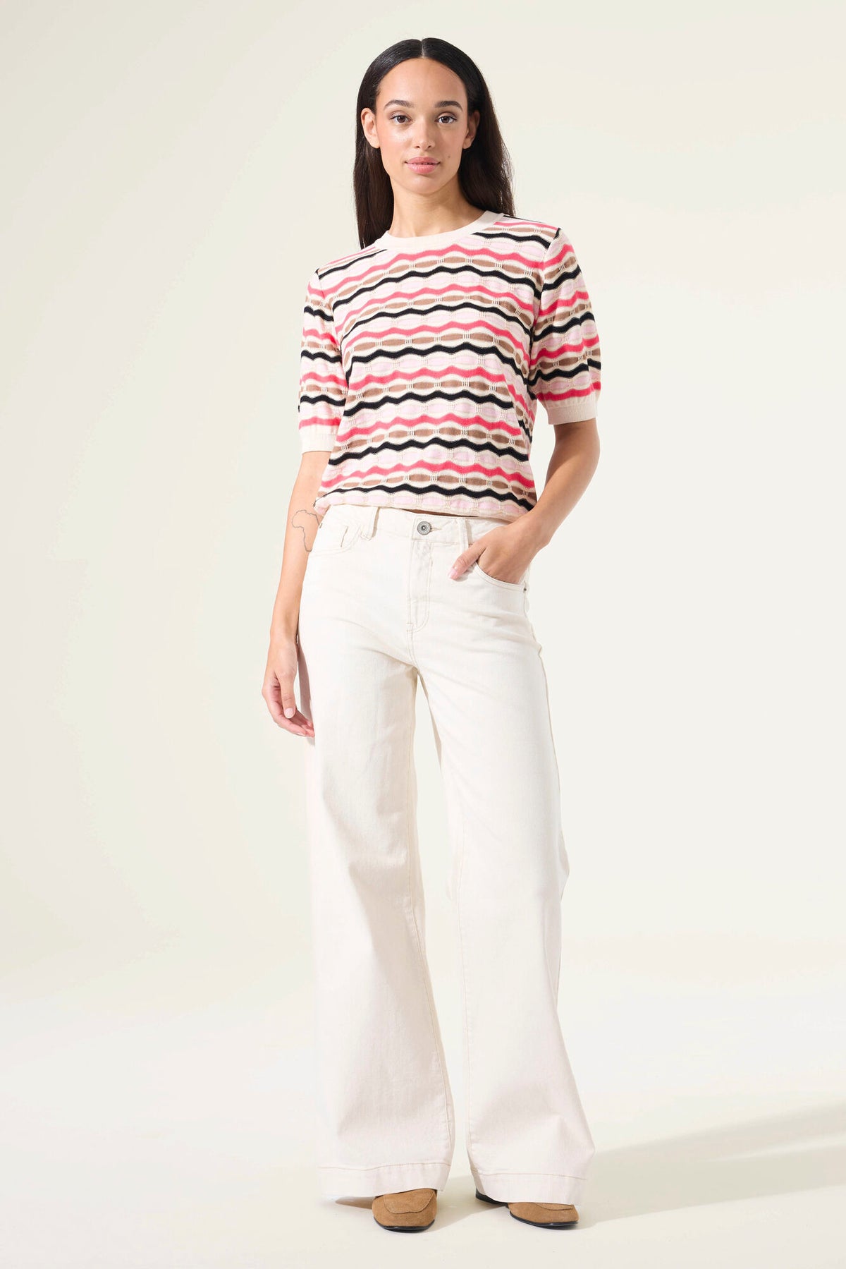 Woman wearing a short sleeve striped sweater and white pants against a plain background
