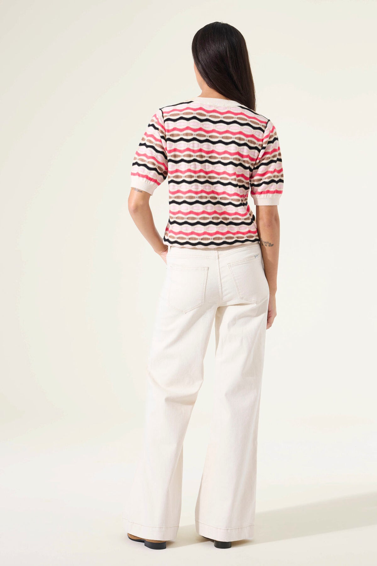 Woman shown from the back wearing a short sleeve striped sweater and white pants against a plain background