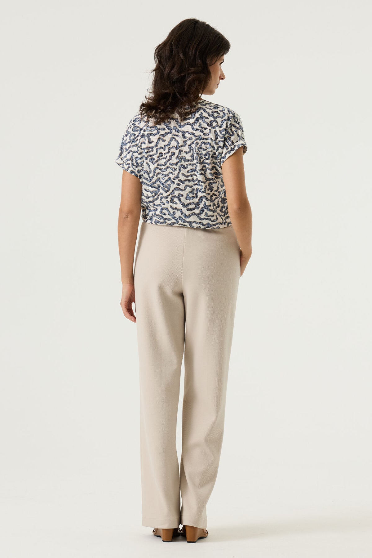woman shown from the back wearing a white v neck t-shirt with blue pattern  and cream pants on a plain background