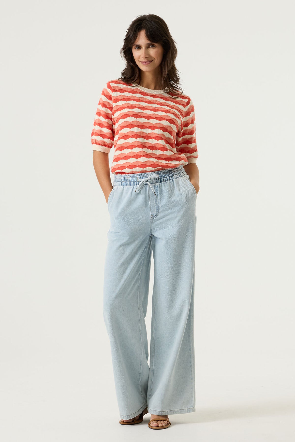 Woman wearing a red and white patterned short sleeve sweater and light wide leg denim pants on a plain background
