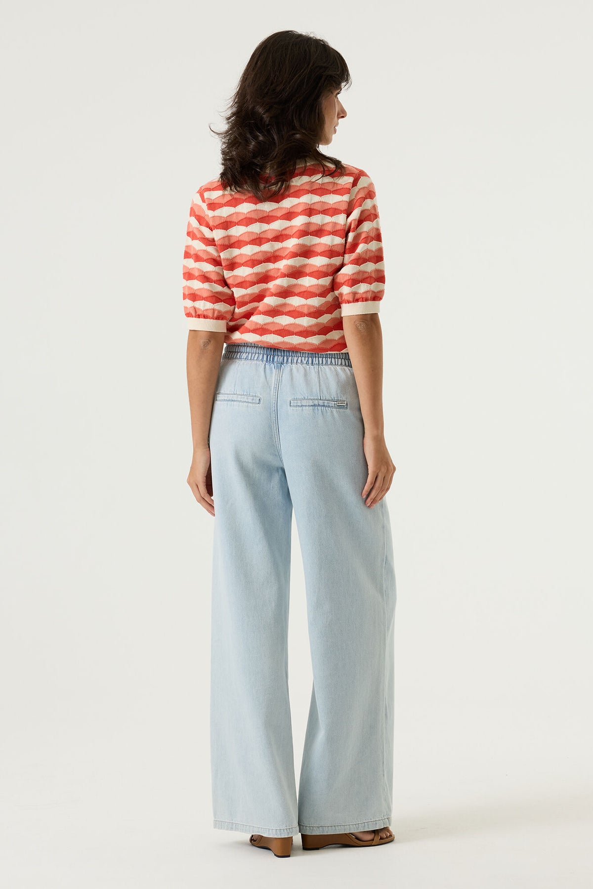 Woman shown from the back wearing a red and white patterned short sleeve sweater and light wide leg denim pants on a plain background