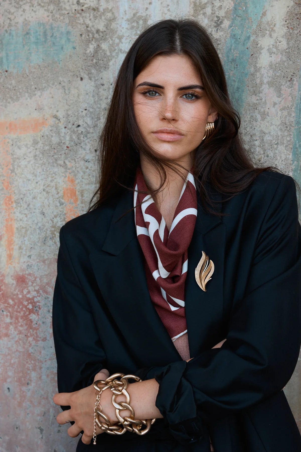 Woman wearing a dark coat and patterned pinot and white colour scarf against a textured wall.