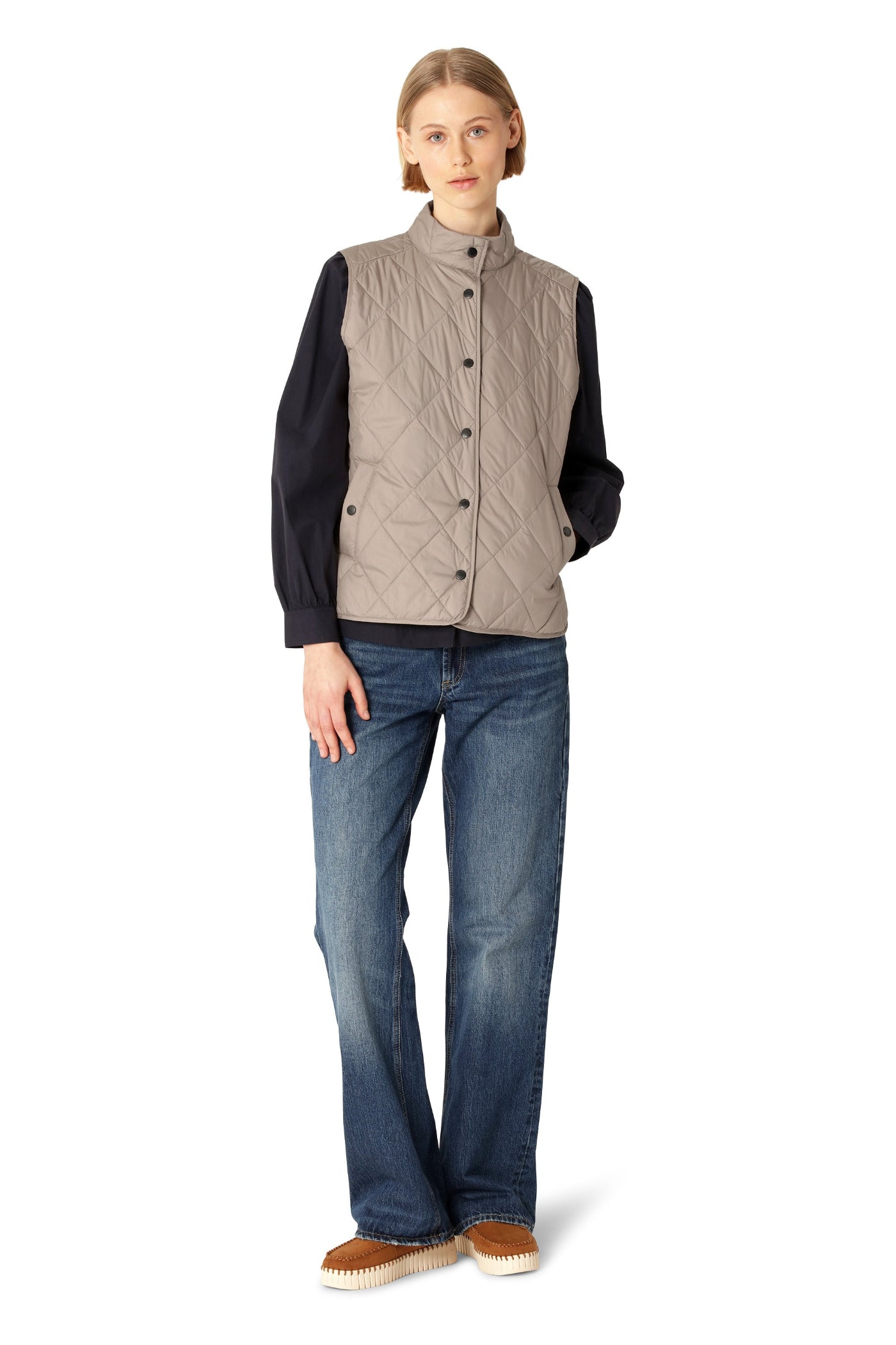 Woman wearing a beige quilted vest over a black shirt with blue jeans on a white background