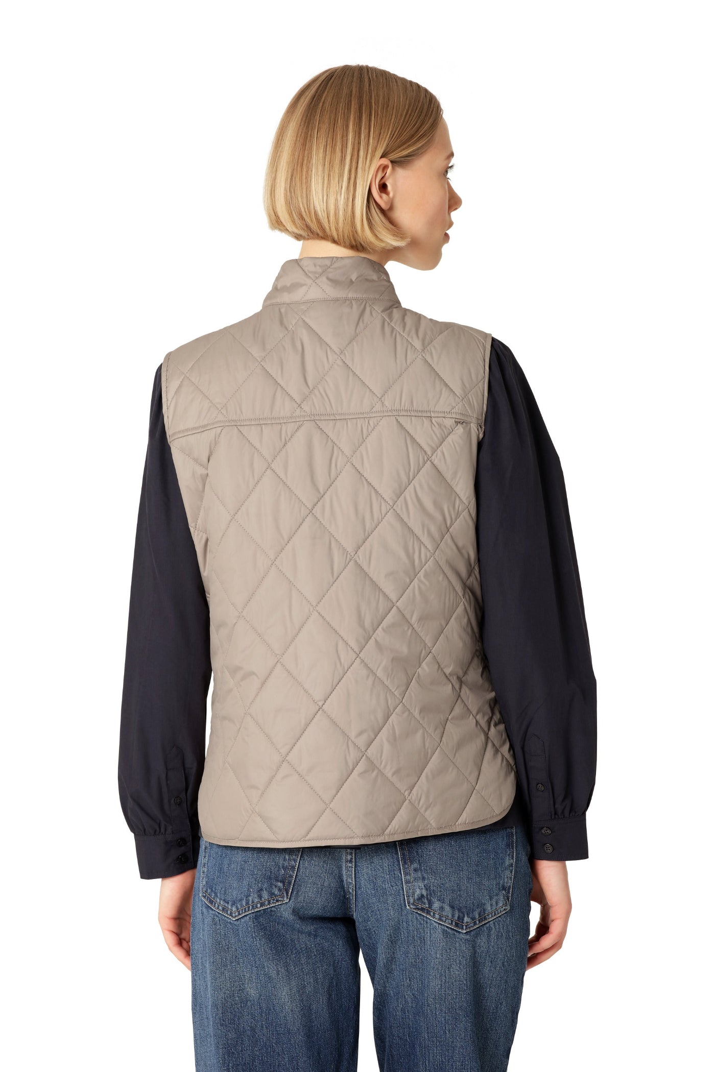 Person wearing a beige quilted vest over a dark long-sleeve shirt and blue jeans on a white background