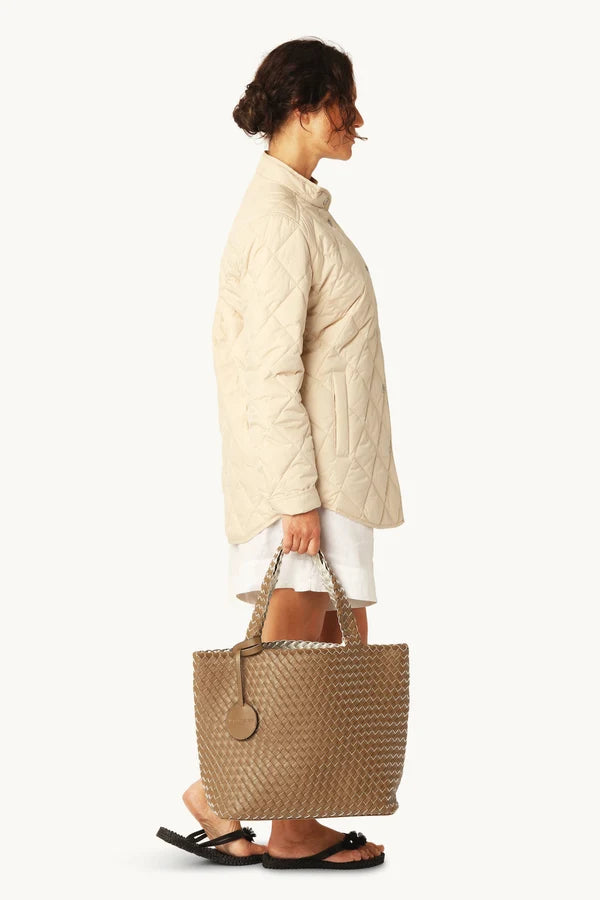 Woman holding a woven handbag wearing a beige quilted jacket on a white background