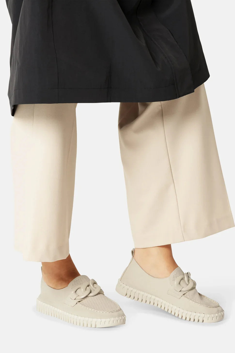 A close-up of a person's lower legs wearing beige knit slip-on loafers with decorative  details styled with beige cropped trousers and a black blazer against a light background.
