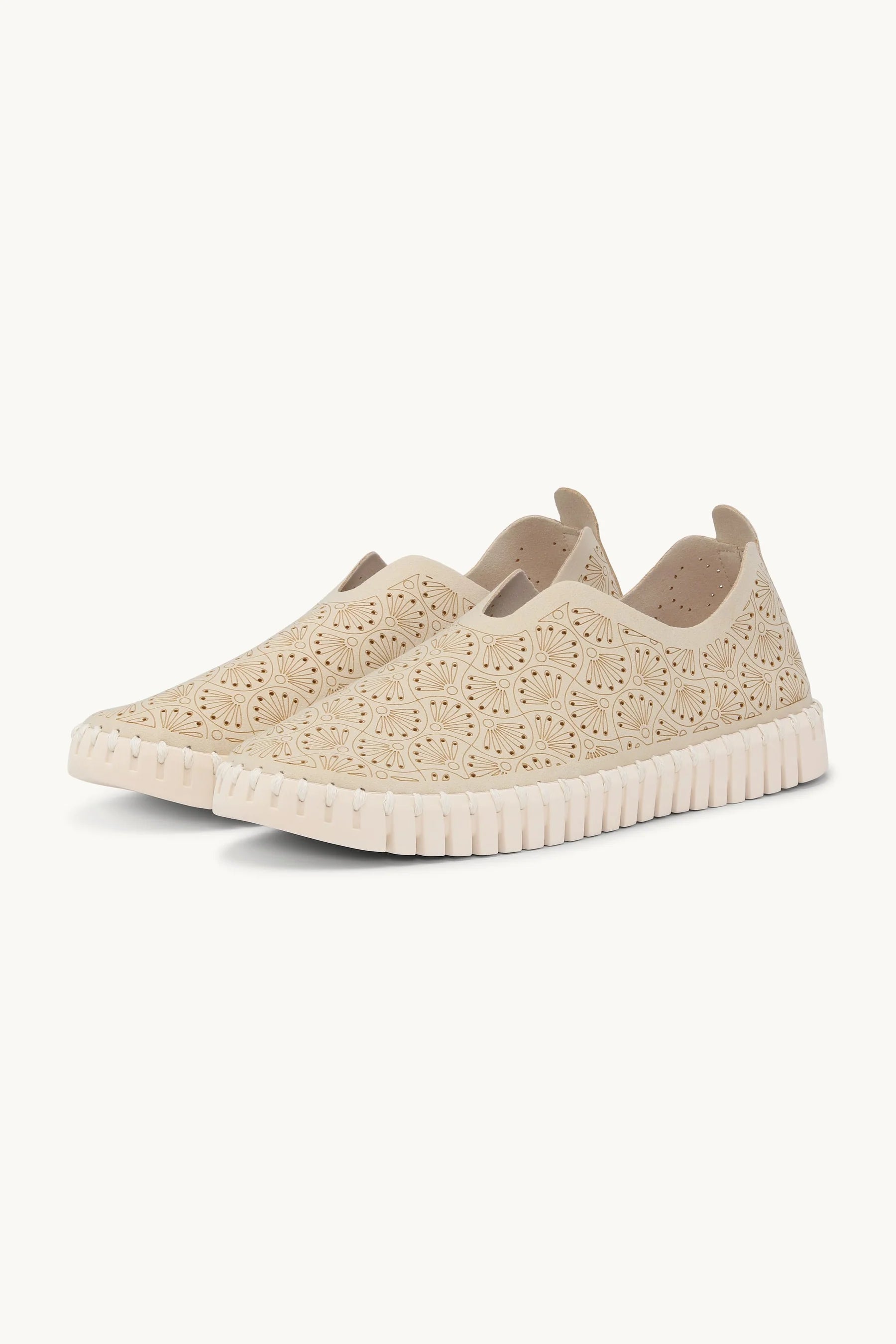 A pair of beige slip-on sneakers with intricate floral cutout embroidery on the upper and a textured, wavy sole, shown against a plain off-white background.