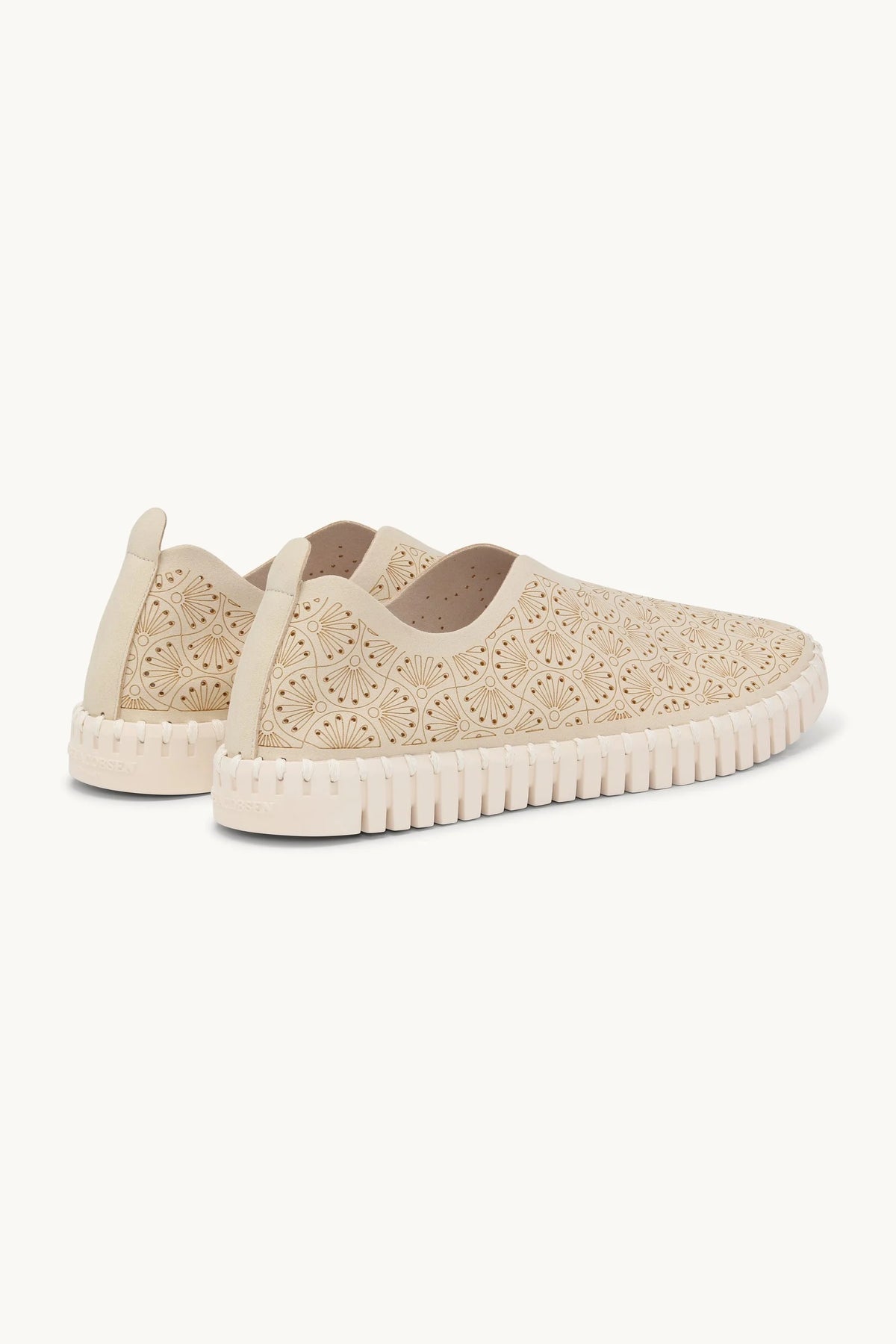A pair of beige slip-on sneakers with intricate floral cutout embroidery on the upper and a textured, wavy sole, shown against a plain off-white background.