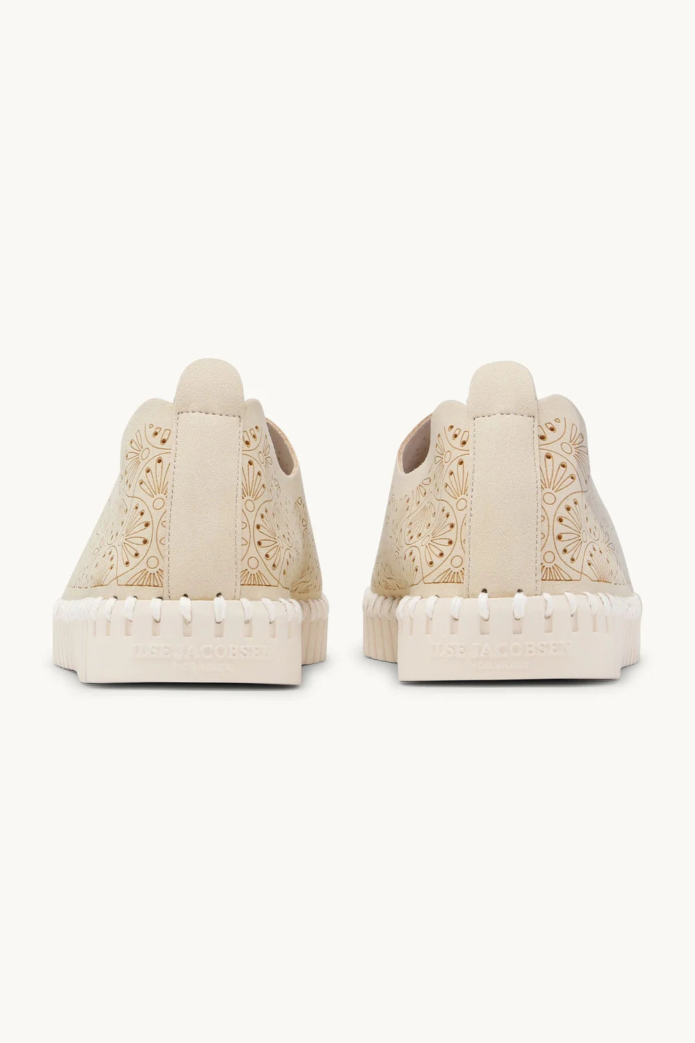 A pair of beige slip-on sneakers with intricate floral cutout embroidery on the upper and a textured, wavy sole, shown against a plain off-white background.