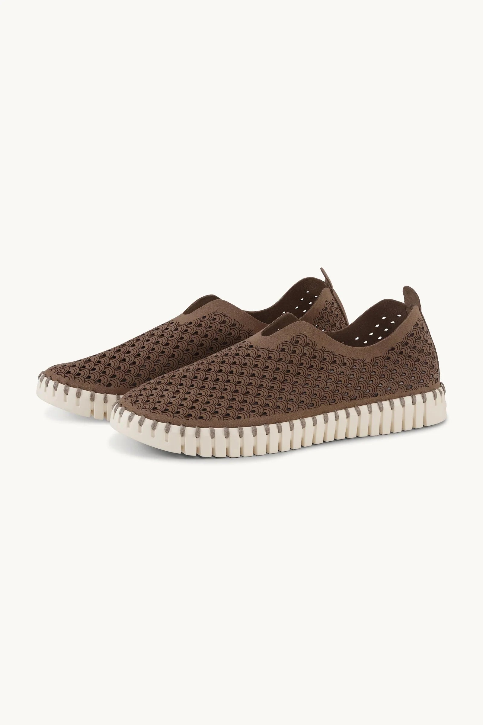 Pair of brown slip on shoes with lazer cut patter and white rubber soles on a plain background. 