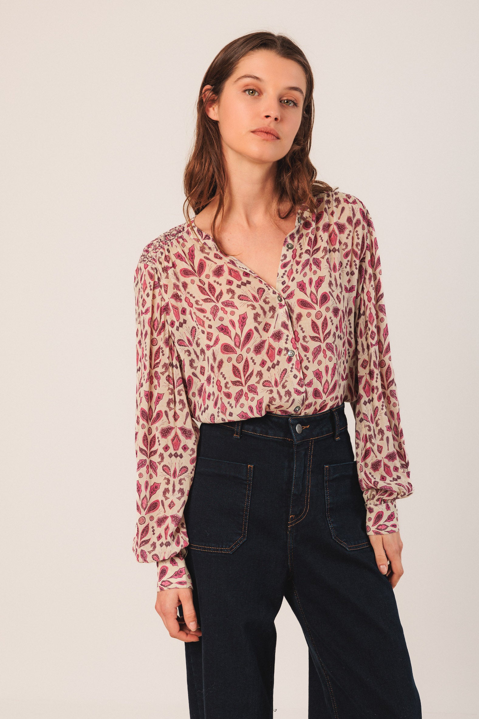 Woman wearing a patterned blouse and dark jeans on a plain background