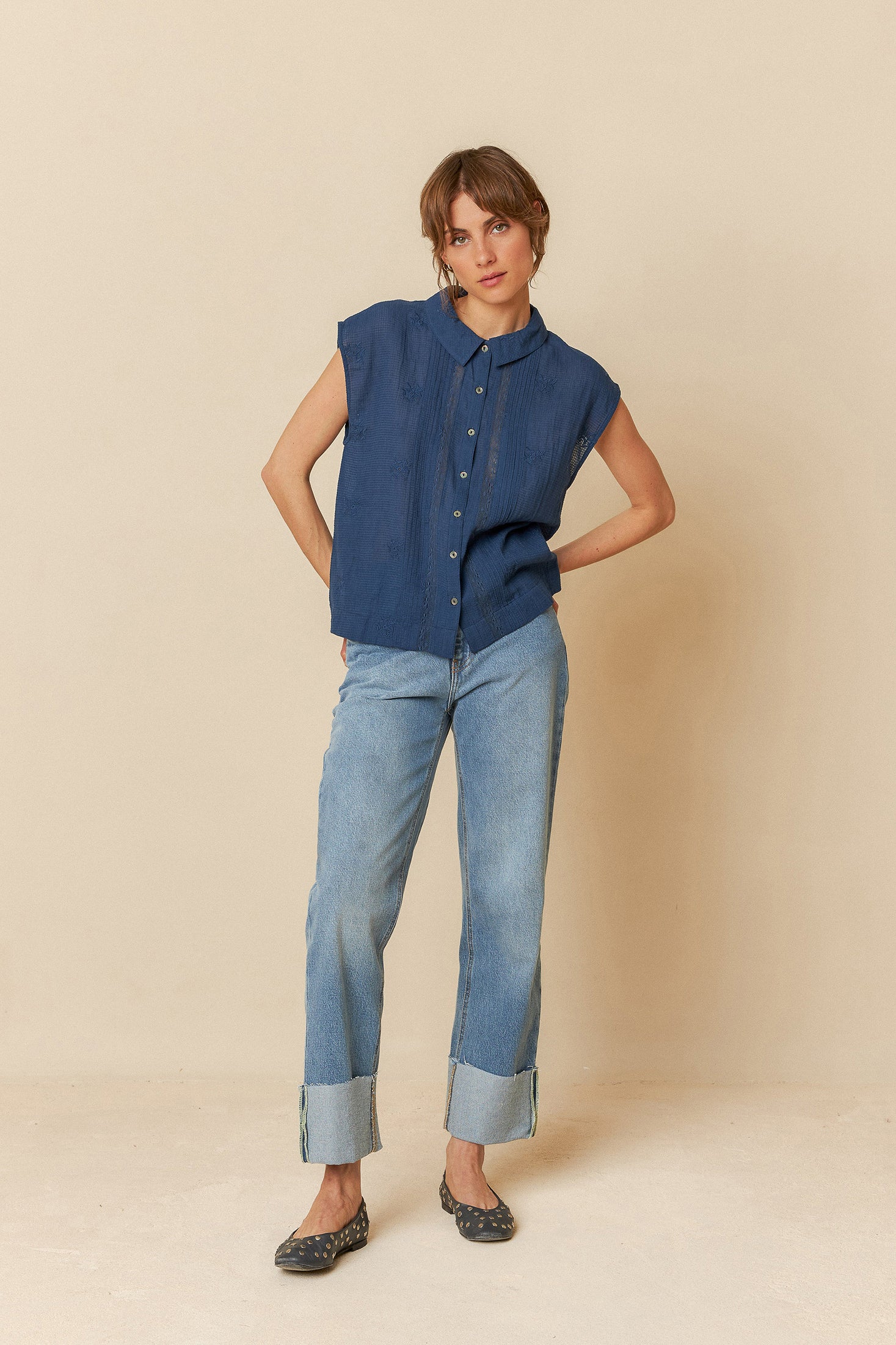 Model wearing a blue sleeveless blouse denim jeans on a beige background. 