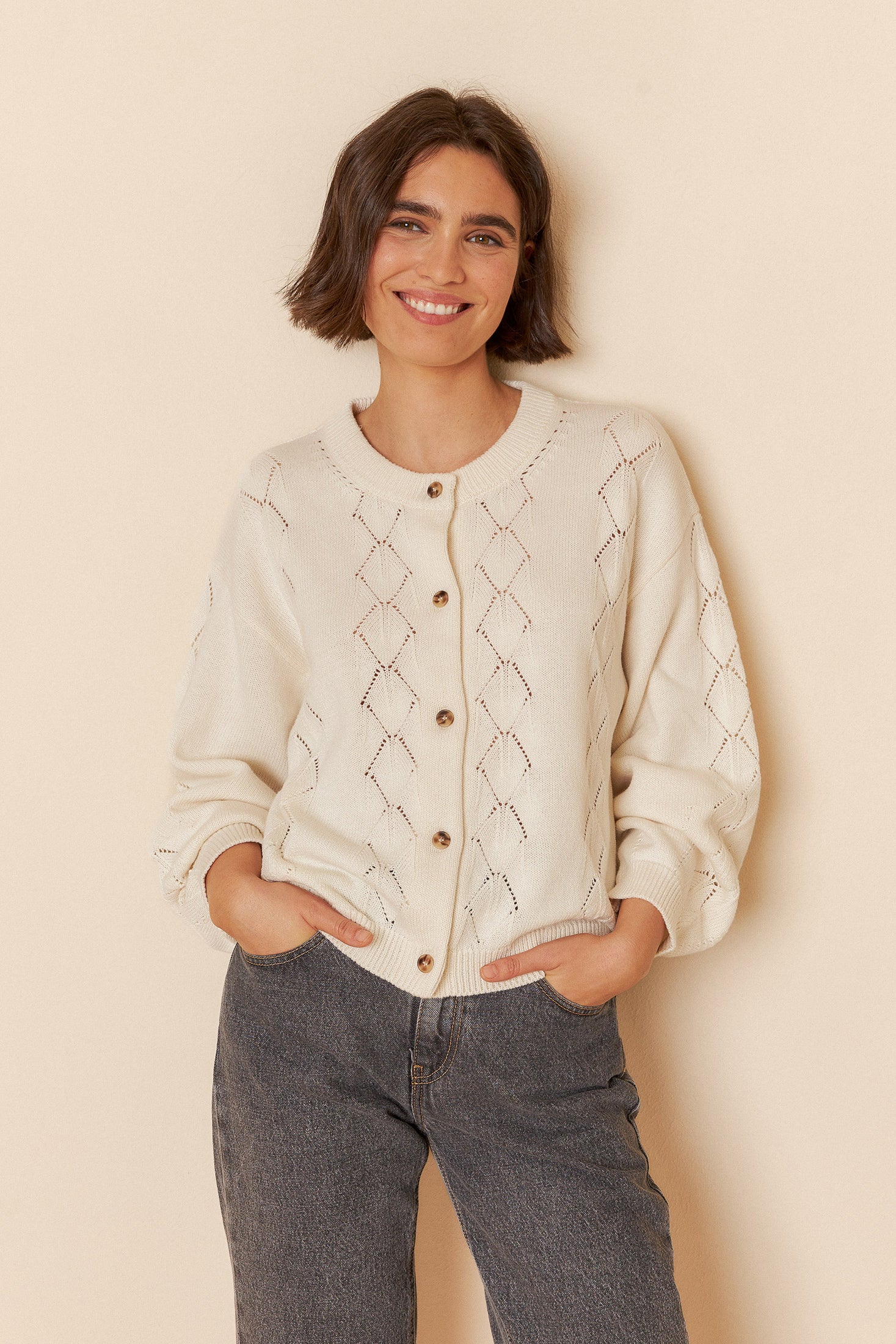Model wearing a white sweater with geometric patterns and grey jeans on a beige background
