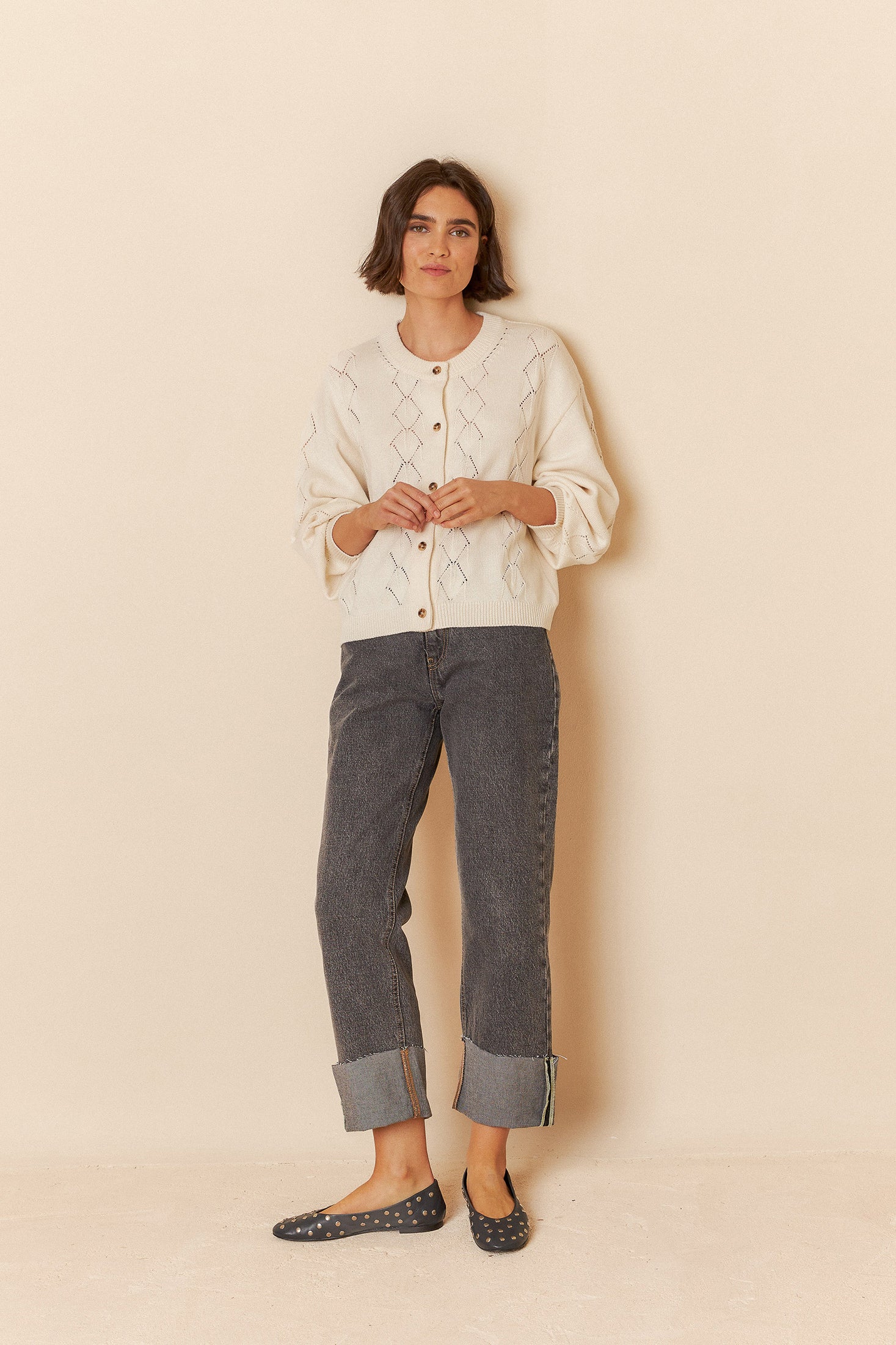 Model wearing a white sweater with geometric patterns and grey jeans on a beige background