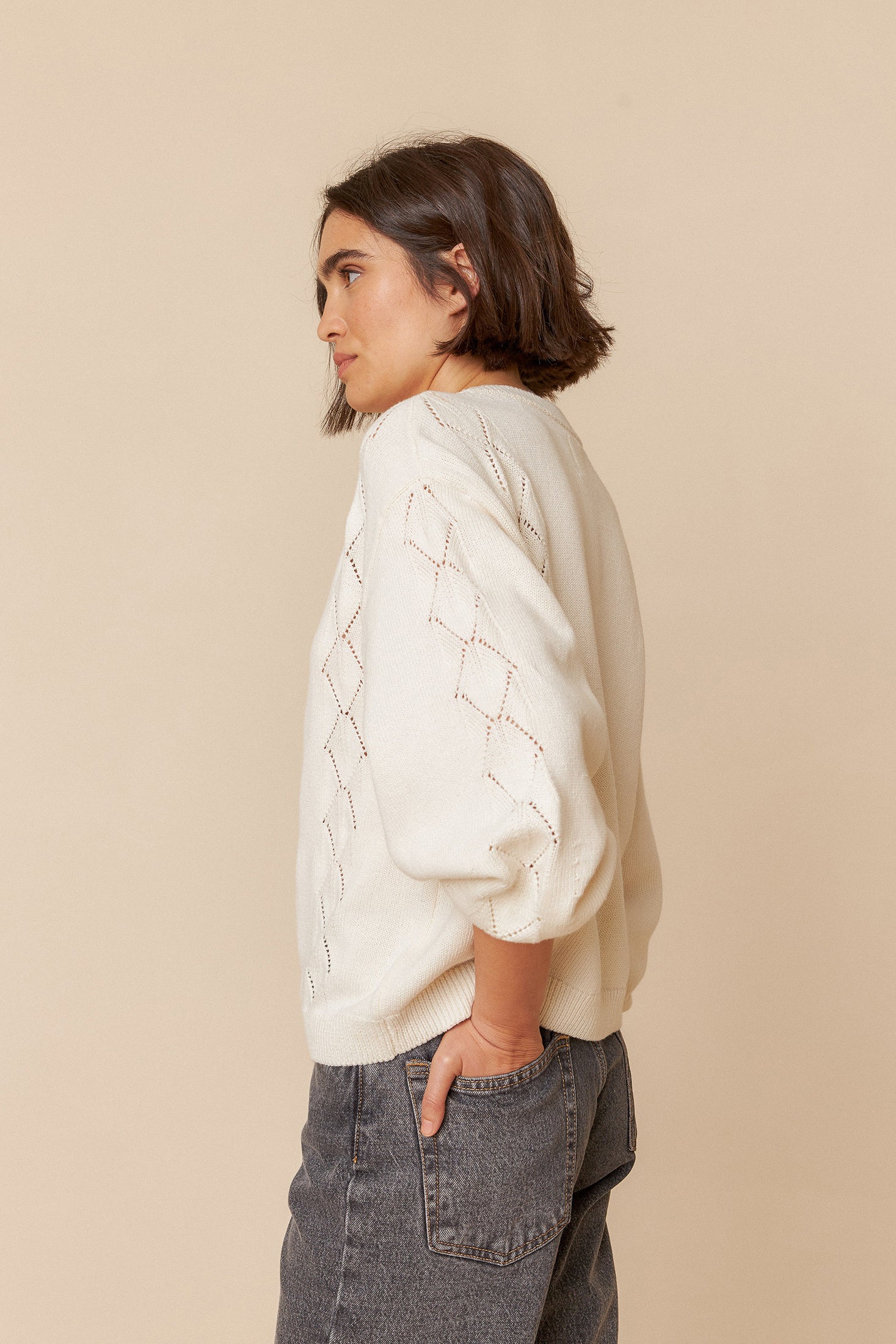 Model wearing a white sweater with geometric patterns and grey jeans on a beige background