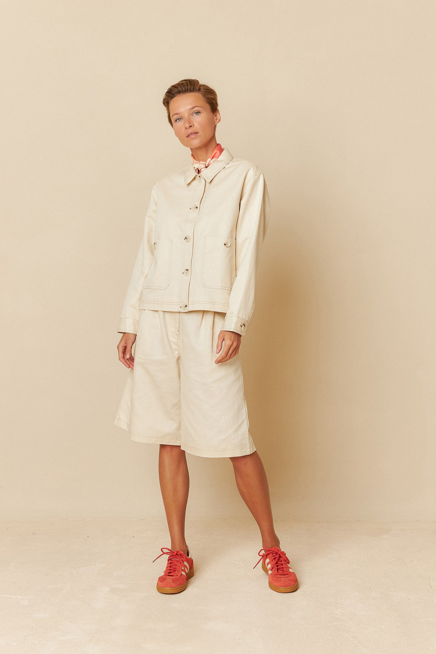 Woman wearing a green and white top  and ecru bemuda shorts standing in front of a colourful wall