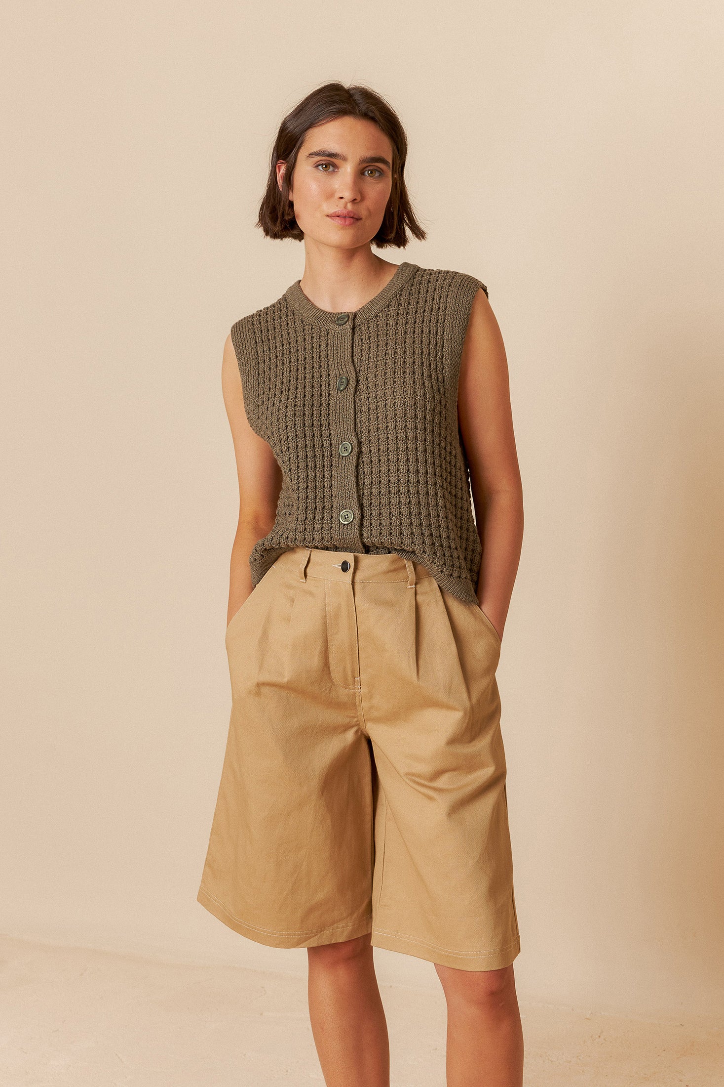 Woman wearing a sleeveless green top and beige bermuda shorts against a beige background